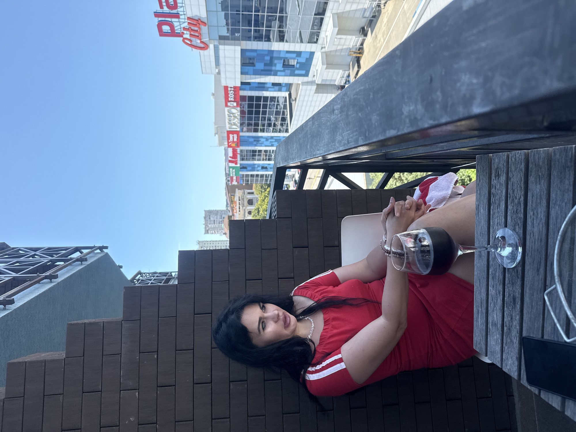 A woman sits on a balcony overlooking a cityscape. She has dark hair and is wearing a red dress. In front of her is a glass of red wine and a cell phone on a wooden table. The buildings in the background appear to be commercial structures with signage. The sky is clear and blue. The image appears to have been taken in Sochi, Russia, as indicated by the "Plaza City" sign visible on one of the buildings. The overall scene is one of leisure and relaxation on a sunny day.