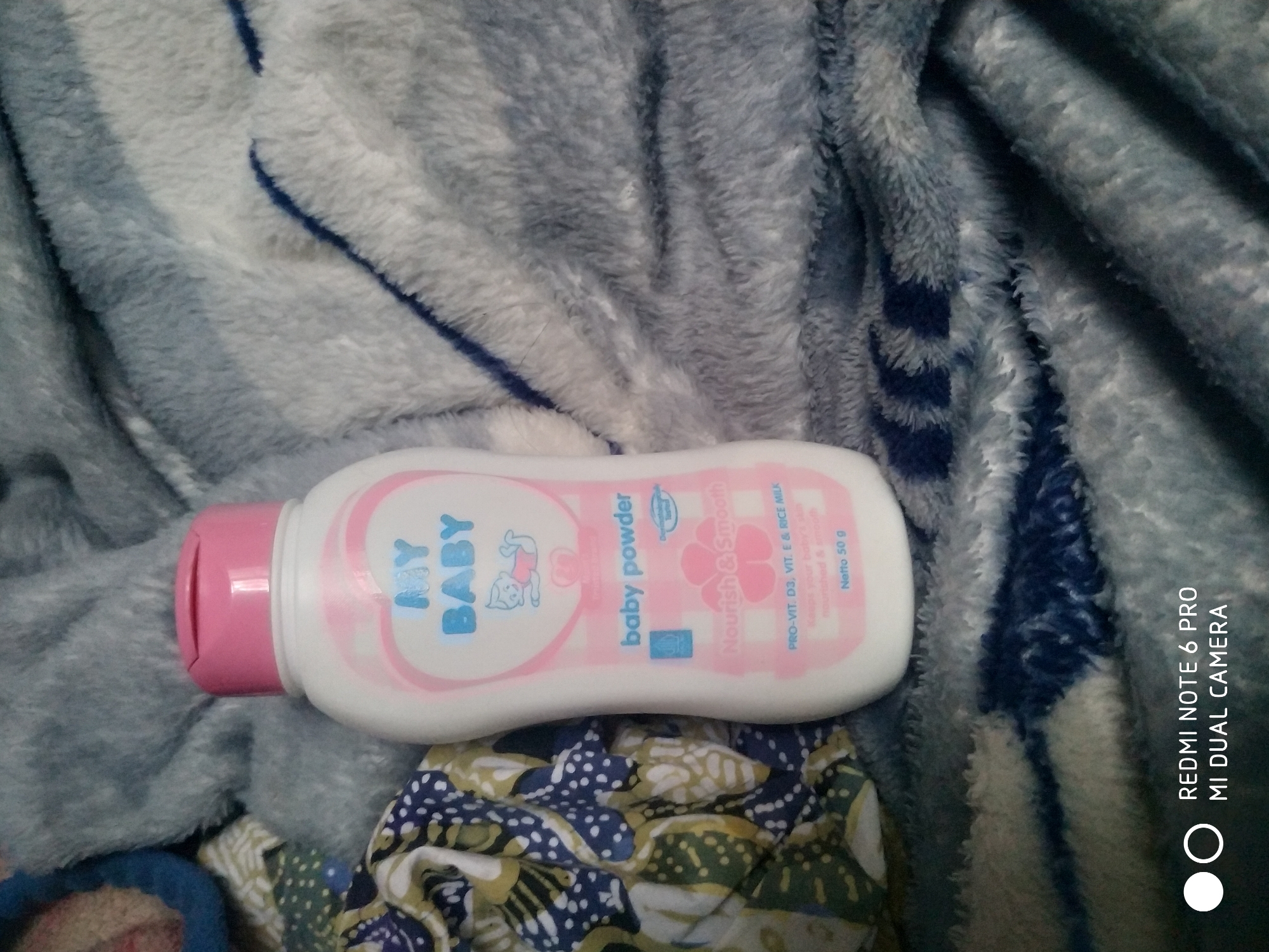 A white bottle of "MY BABY" brand baby powder with a pink cap sits on a soft, textured gray blanket. The bottle features blue text that reads "MY BABY" and a drawing of a baby. Below that, it says "baby powder" in blue. Further down the bottle, pink text indicates "Nourish & Smooth" and lists "PRO-VIT. D3, VIT. E & RICE MILK" and "Netto 50 g". To the left of the bottle, a portion of a colorful fabric with a blue and white patterned design is visible. The image is framed by the same gray blanket material. The scene is not indicative of any specific location within the city of Badas, Indonesia.