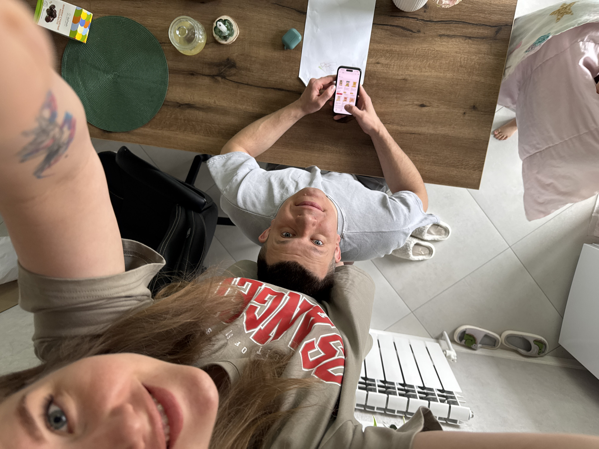 A couple is taking a selfie indoors. The man is sitting at a wooden table, looking at a smartphone. The woman is holding the camera, her face close to the lens. On the table are a round green placemat, a small box, two small glass containers, a white sheet of paper, and a blue object. The man is wearing a gray t-shirt. The woman is wearing a gray sweatshirt with red letters. A black backpack is visible near the man. A white bedspread with stars is on the right side of the table. White tile flooring is present. A white radiator is visible in the lower right. The location is presumed to be indoors in the city of Gornyi (Dvurechenskoe S/P), Russia.