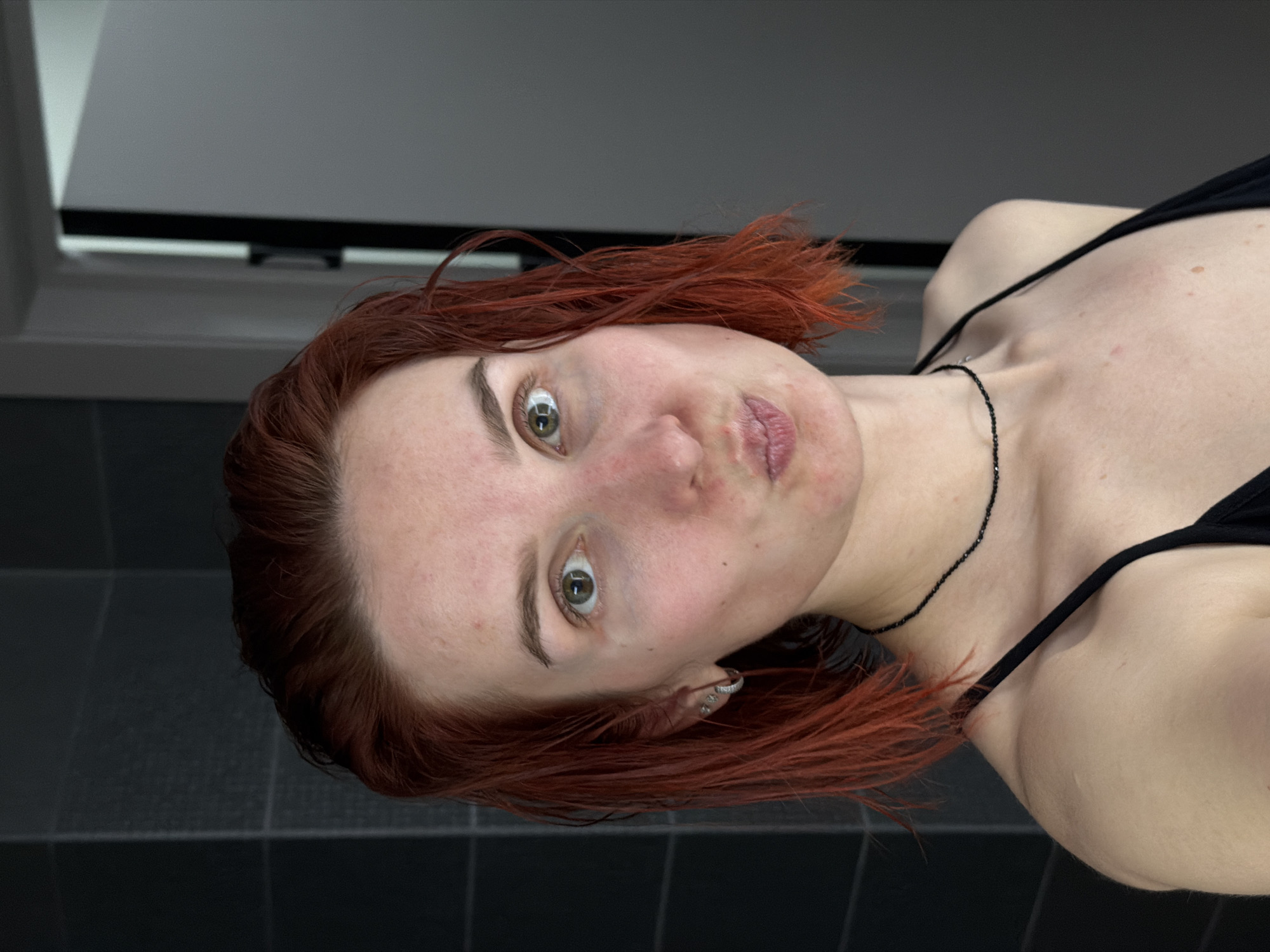 A woman with auburn hair, wearing a black top and a black beaded necklace, is lying on a black tiled surface in what appears to be a bathroom. She has light skin, green eyes, and a pierced ear. The background includes a white wall above dark tiled flooring. The image was most likely taken inside a bathroom. There is no information to indicate that this image was taken in the city of Chaikovsky, Russia.