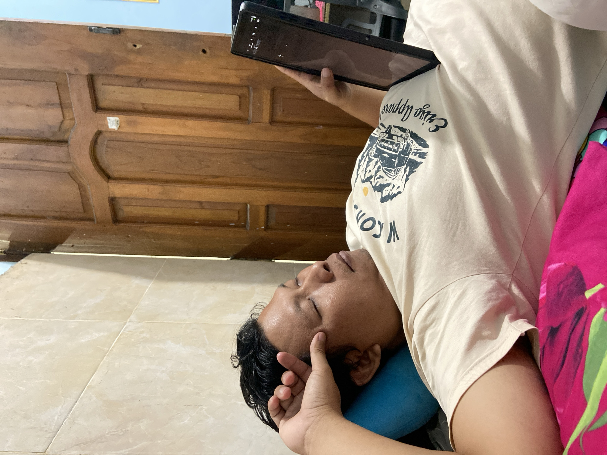 The image captures a man lying on a light-colored tiled floor indoors, with his head resting on a blue cushion or pillow. His eyes are closed or partially closed, and one hand is gently placed on his temple, suggesting relaxation or rest. With his other hand, he holds a black tablet or smartphone, which has a dark screen displaying faint user interface elements.
He is wearing a light beige or cream-colored t-shirt with a dark graphic print on the chest. Visible text on the shirt includes "Original Apparel" along with a design that appears to be a vintage car or van. Next to him, a vibrant pink and green patterned fabric, possibly a blanket or bedspread, is visible.
A wooden door with horizontal paneling and decorative elements dominates the background, indicating an indoor, likely domestic, setting. The lighting suggests it is daytime. The scene conveys a sense of calm and leisure in what is likely a home in Gelam, Indonesia.