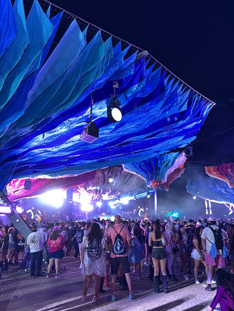 A nighttime outdoor crowd gathers under large, blue, tent-like structures with suspended lighting fixtures. The structures appear to provide shade or decoration above the crowd. The crowd is diverse, and many people are wearing casual clothing. The crowd is focused towards a bright stage area in the distance, suggesting a performance or event is taking place. The setting appears to be at Coachella, in Indio, United States, due to the style of the outdoor event, the large tent-like structures used for decoration, and the time of day.