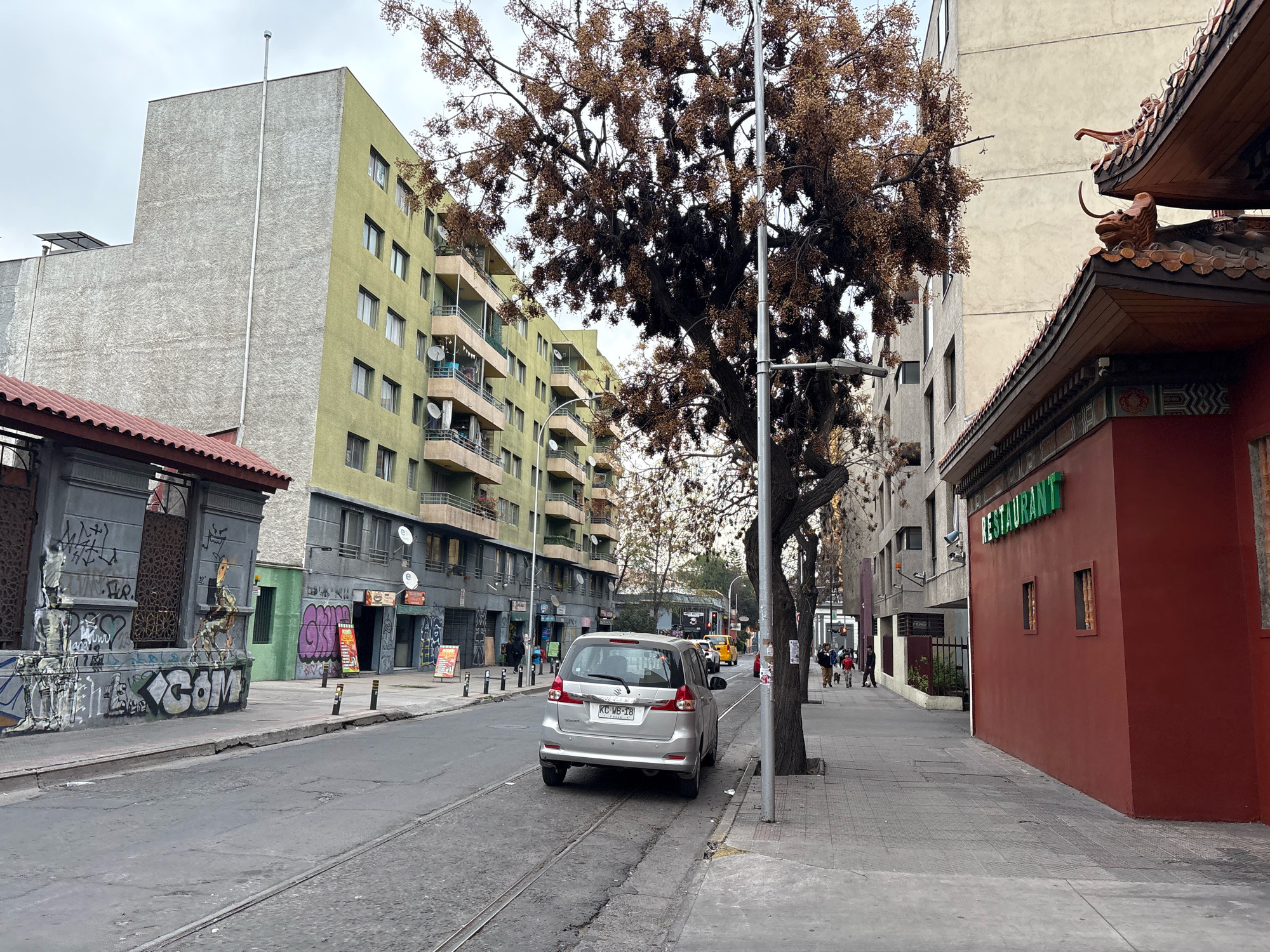A silver Suzuki Ertiga is parked on a city street in Santiago, Chile, with tram tracks running in the road. The vehicle's license plate is visible as KC WB-18. To the left of the car, a building with a textured gray facade and lime green accents stands, featuring balconies with satellite dishes and clotheslines. Across the street, on the right, a red building with a traditional Chinese architectural style roof is situated, with a green sign that reads "RESTAURANT" above it. Further down the street, more buildings of various heights and colors line the sidewalk. Several pedestrians are walking on the sidewalk, and a yellow taxi is visible in the distance. A large tree with sparse brown leaves is in the foreground, obscuring part of the view. Graffiti adorns a wall on the far left of the image. The sky is overcast.