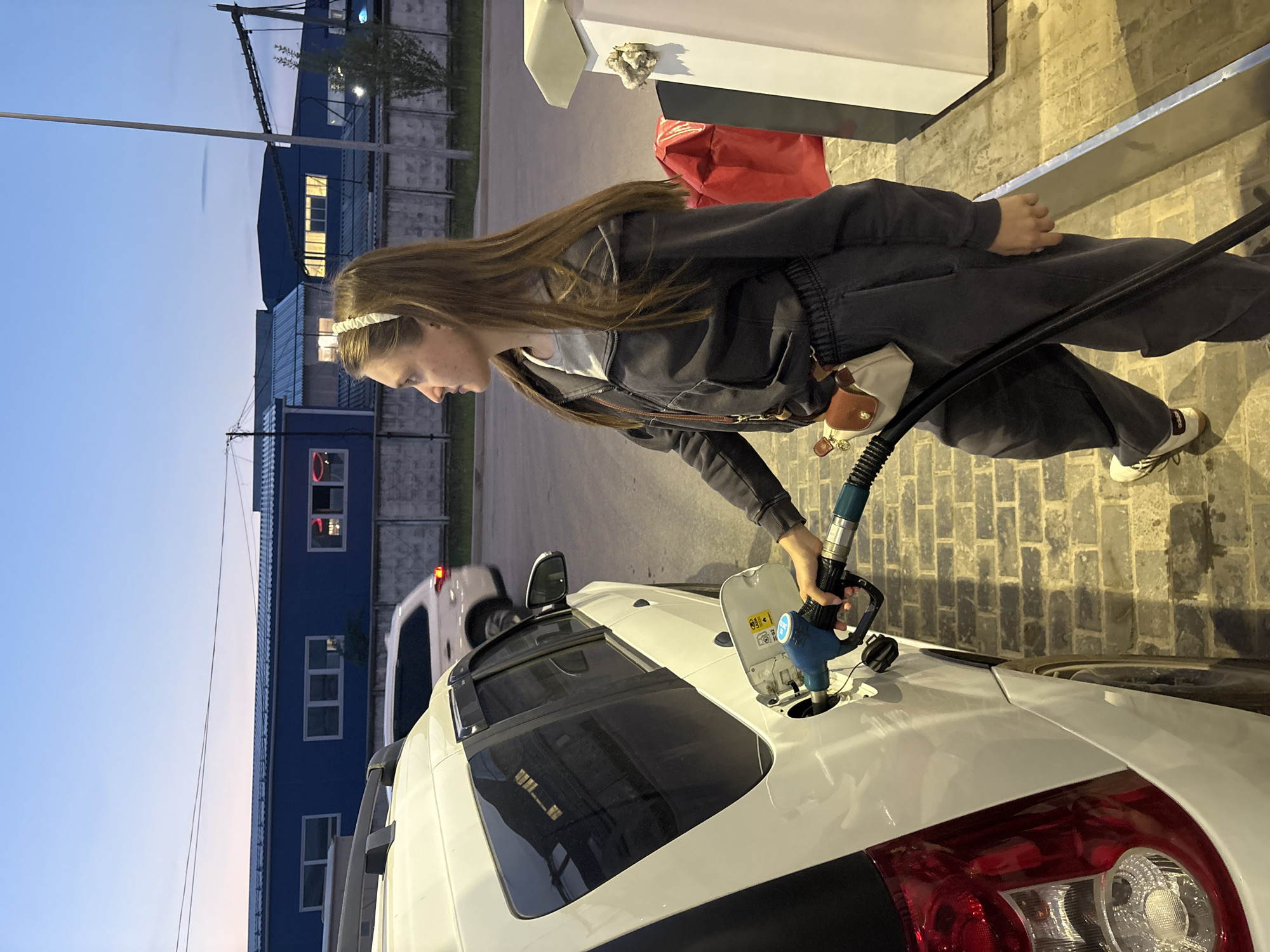 A woman is filling the gas tank of a white car at a gas station. The car is parked next to a gas pump. The woman is wearing a dark sweatsuit and a white headband. She has long brown hair. The building across the street is blue and white. The time of day appears to be early evening, with a dusk sky. The location is Perm, Russia.