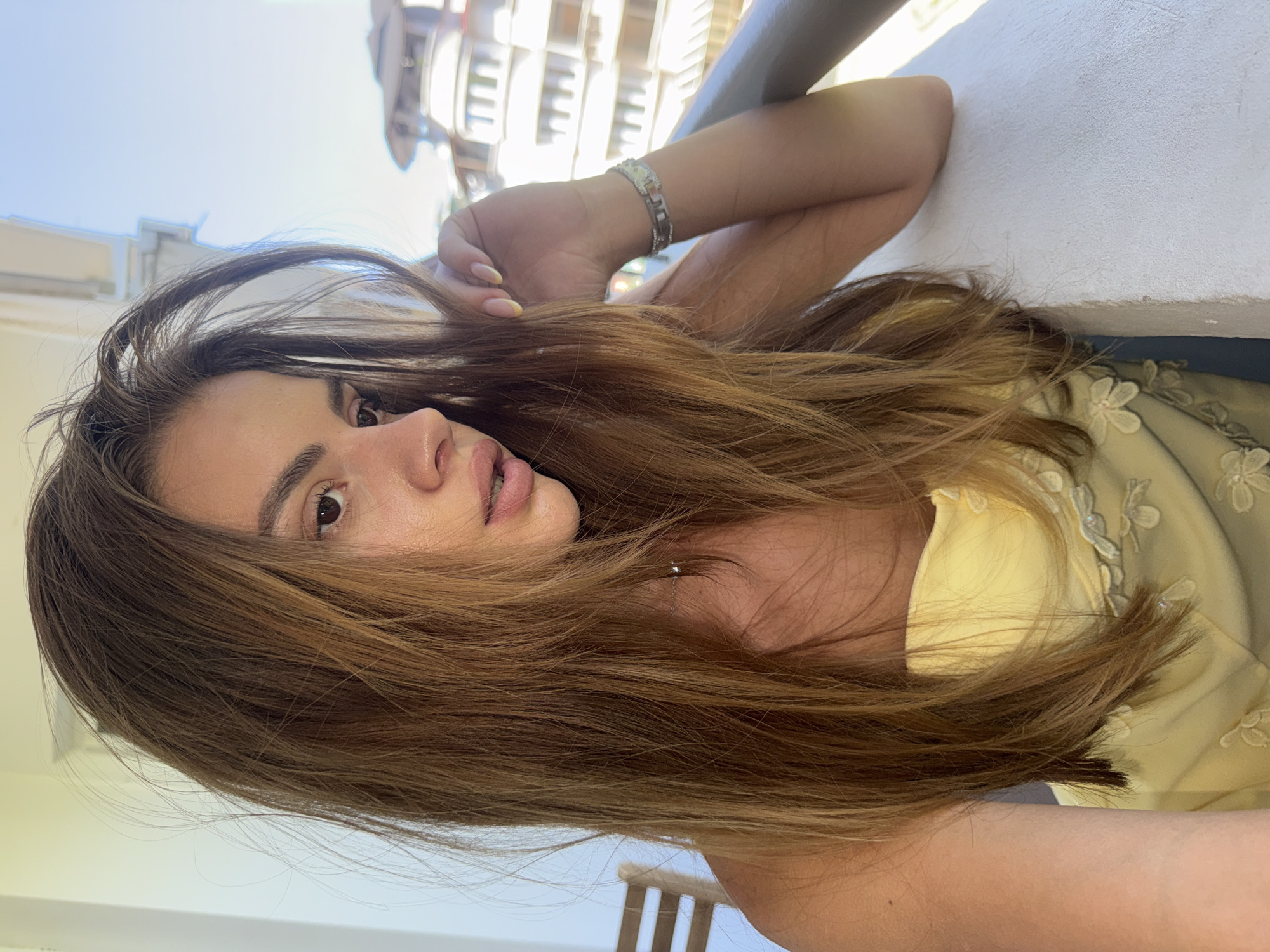 A woman with long, brown hair and light skin is looking down and to the right, with her right arm resting on a white railing. She is wearing a yellow strapless top with floral embellishments. Her left hand is touching her hair. In the background, there is a blurry building with white balconies and a clear blue sky. The image appears to be taken outdoors on a sunny day, possibly on a balcony in Plovdiv, Bulgaria, given the architectural style of the background building.