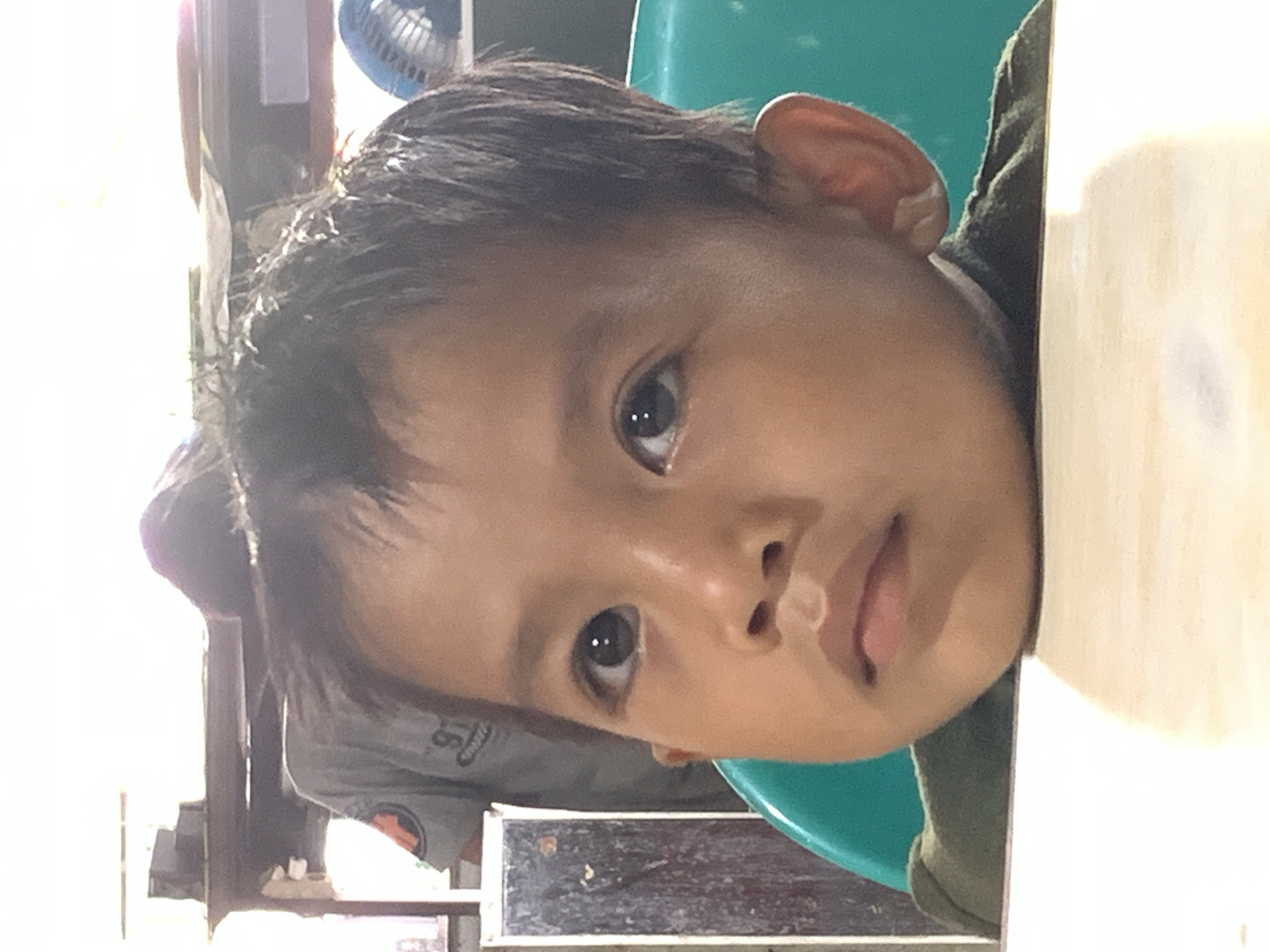 A close-up portrait of a young child with dark hair and eyes, resting their chin on a white surface, possibly a table. The child is wearing a dark green shirt. Behind the child, a blue plastic chair is visible. In the blurred background, parts of a room setting can be discerned, with an unidentified person wearing a grey shirt with what appears to be an emblem. There's also a glimpse of a fan. The exact location in Samarinda, Indonesia, cannot be determined from the image.