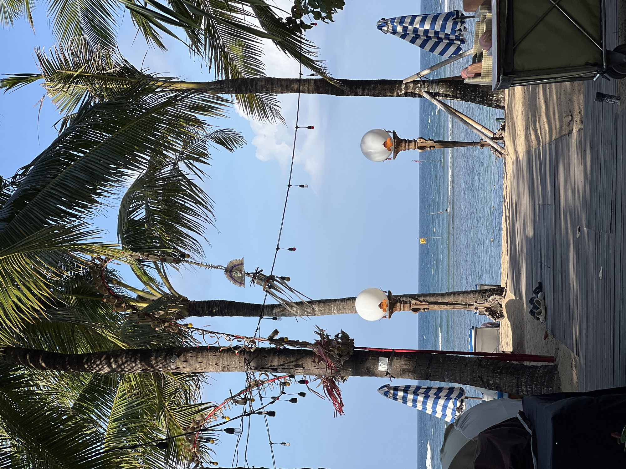 This media file captures a vibrant beach scene in Denpasar, Indonesia. The primary subjects are palm trees with their fronds arching towards the bright blue sky, interspersed with a string of festive lights. A wooden walkway runs along the sandy beach, leading to the ocean where gentle waves roll in. Two blue and white striped umbrellas are visible, suggesting a place of relaxation or a beachside establishment.
There are no people actively engaged in activities, but elements like a lounge chair with a person's legs visible, and some footwear on the walkway, hint at recent or ongoing human presence. Decorative objects, including what appear to be traditional Indonesian adornments hanging from the palm trees and a rustic lamp post with a large globe, add character to the scene.
The time of day appears to be late morning or early afternoon, indicated by the bright sunlight casting shadows and the clear, sunny sky with a few scattered clouds. The weather seems pleasant and warm, typical of a tropical beach. The overall mood is tranquil and inviting, evoking a sense of vacation or leisure. The location cues like palm trees, beach, and umbrellas strongly point to a tropical coastal setting. No visible text is present in the media file.