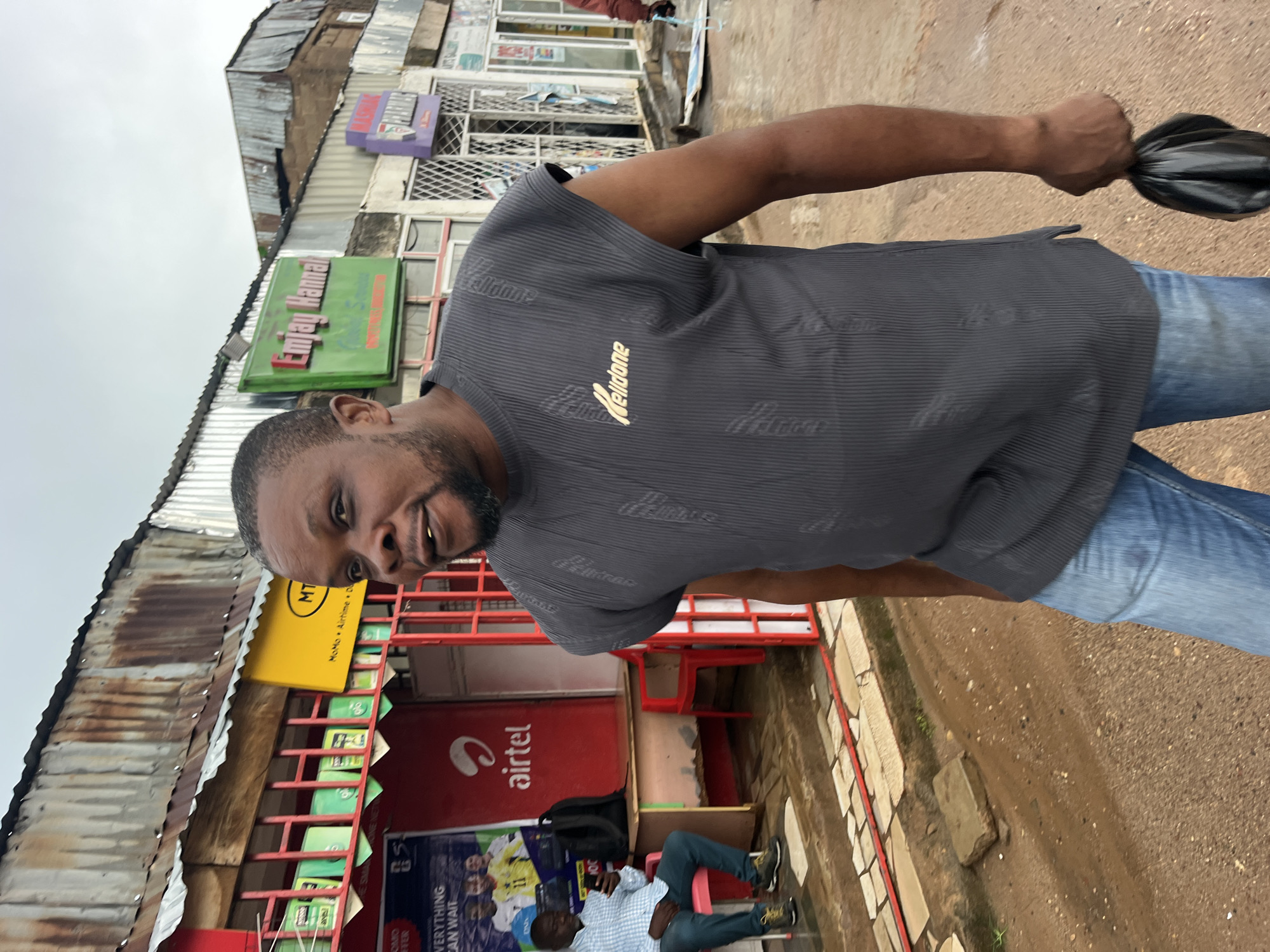 In a street scene, a man in a grey, ribbed t-shirt and blue jeans holds a black plastic bag in his right hand. He has a beard and appears to be looking at the camera. Behind him, a red storefront is visible, with an "Airtel" logo on the red wall. Several other signs are also present, including one in green that reads "Emjay Hanna" and a yellow "MTN MoMo Airtime" sign. The area appears to be a market or a commercial street with multiple shops. Another person is seated on a red chair in front of the store. The ground is a dirt road with some gravel. The sky is overcast, suggesting a cloudy day. The overall impression is of a candid street photograph, possibly taken in an African urban environment, given the prominent "MTN" and "Airtel" branding, which are telecommunication companies operating in Africa.