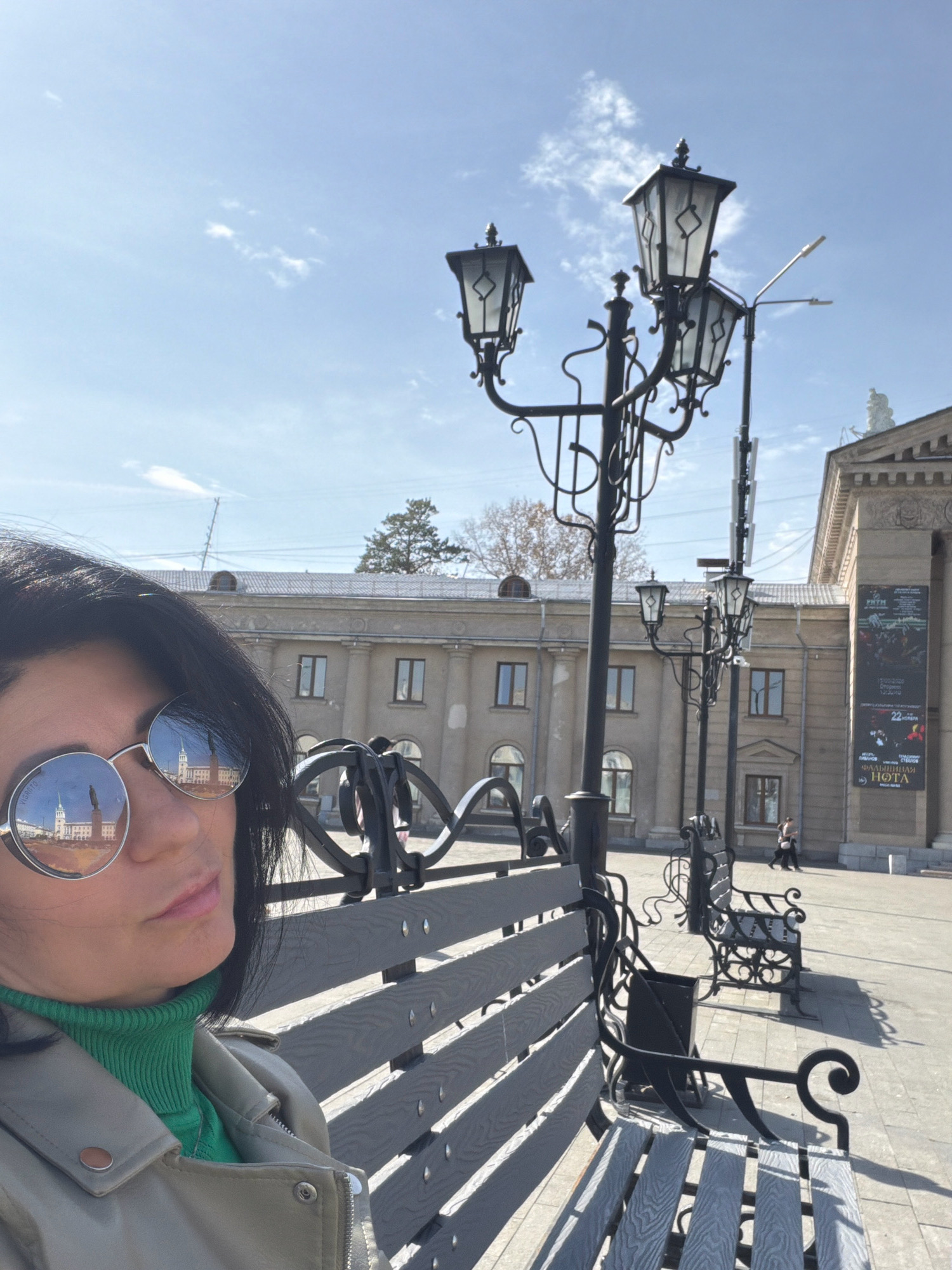 A woman with dark hair and sunglasses is sitting on a park bench in a city square, taking a selfie. The sunglasses reflect a building with columns and a statue. The square has several lampposts and more park benches. In the background, a building with a banner for an event is visible. The environment suggests an urban setting with clear skies. The location is the Angarsk Drama Theater square in Angarsk, Russia.