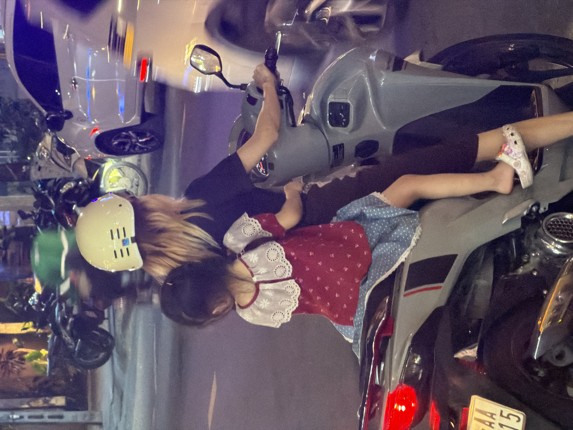 A street scene in Đà Nẵng, Vietnam shows an adult female operating a gray motor scooter in an urban environment at night. She is wearing a black t-shirt and dark pants. A child in a red and white dress and a cream-colored helmet is lying across the scooter’s seat in front of the adult. Other parked motorcycles and a white sedan are also present on the street.