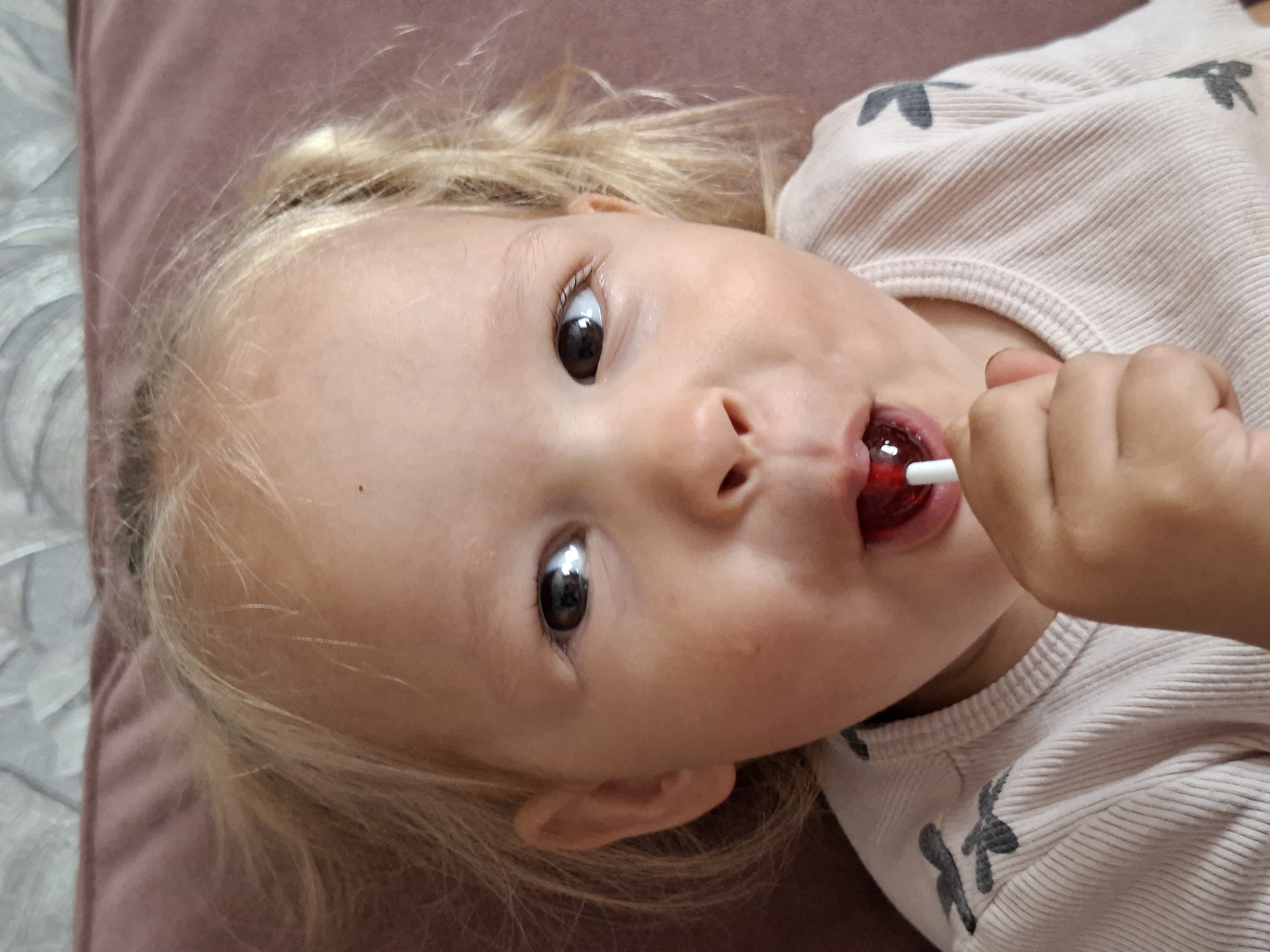 A young child with blonde hair is pictured up close, looking directly at the camera with a surprised expression. They are holding a red lollipop to their mouth. The child is wearing a light-colored, long-sleeved shirt with a subtle ribbed texture and small dark bird-like designs on the shoulder. The background is a soft pinkish-purple surface, likely a sofa or cushion, with a patterned gray fabric visible at the top. The lighting is soft and natural, highlighting the child's face and the texture of their clothing. The image appears to be taken indoors in an unknown urban or suburban setting.