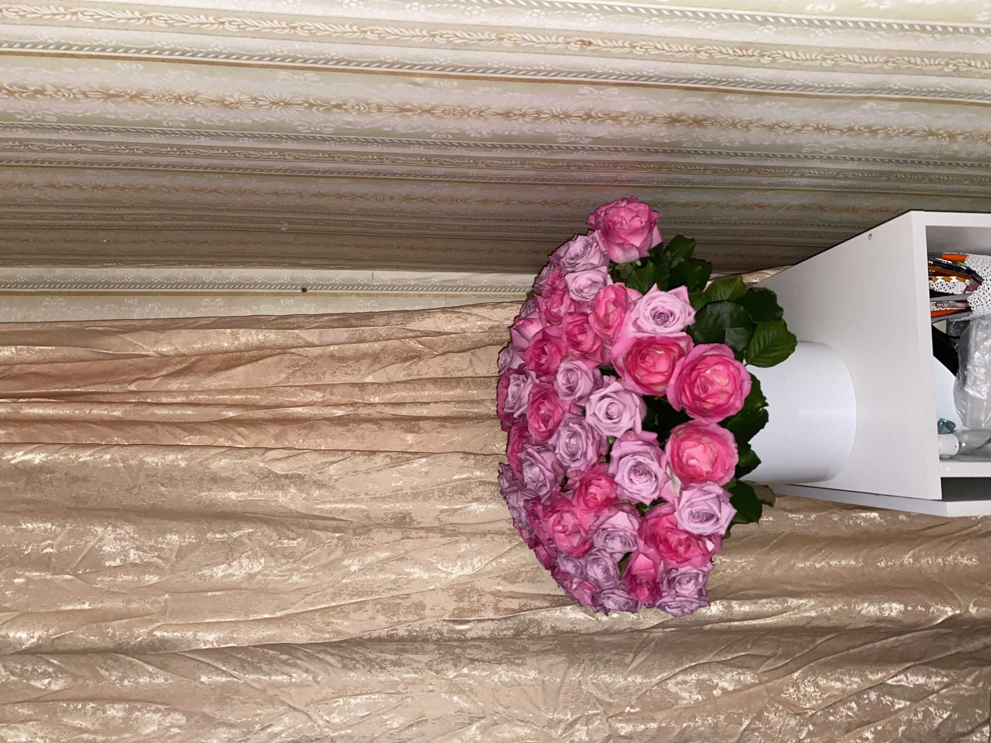A large bouquet of pink and purple roses is displayed in a white vase on a white shelf. The bouquet is positioned in the lower center of the image, against a backdrop of shimmering gold drapes on the left and patterned wallpaper on the right. The shelf also contains a basket of pens and a clear bottle. The scene appears to be indoors.
