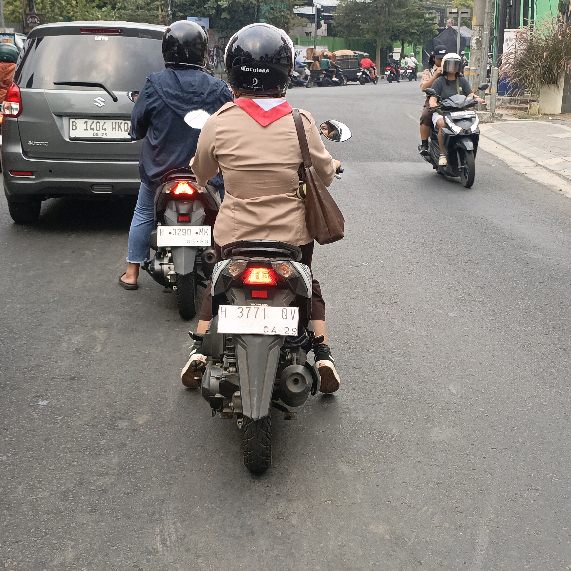 In Salatiga, Indonesia, a dark grey car is parked on the right side of a road. A person wearing a dark blue jacket, blue jeans, and a black helmet is riding a motorcycle behind the car. The motorcycle has a license plate that reads "B 1404 HKQ". Further down the road, another person is riding a motorcycle and wearing a tan uniform with a red and white scarf. This person is also wearing a black helmet. Behind them, another person is riding on the same motorcycle, wearing a dark grey shirt and a white helmet with a visor. The road surface is asphalt. In the background, there are trees and buildings.