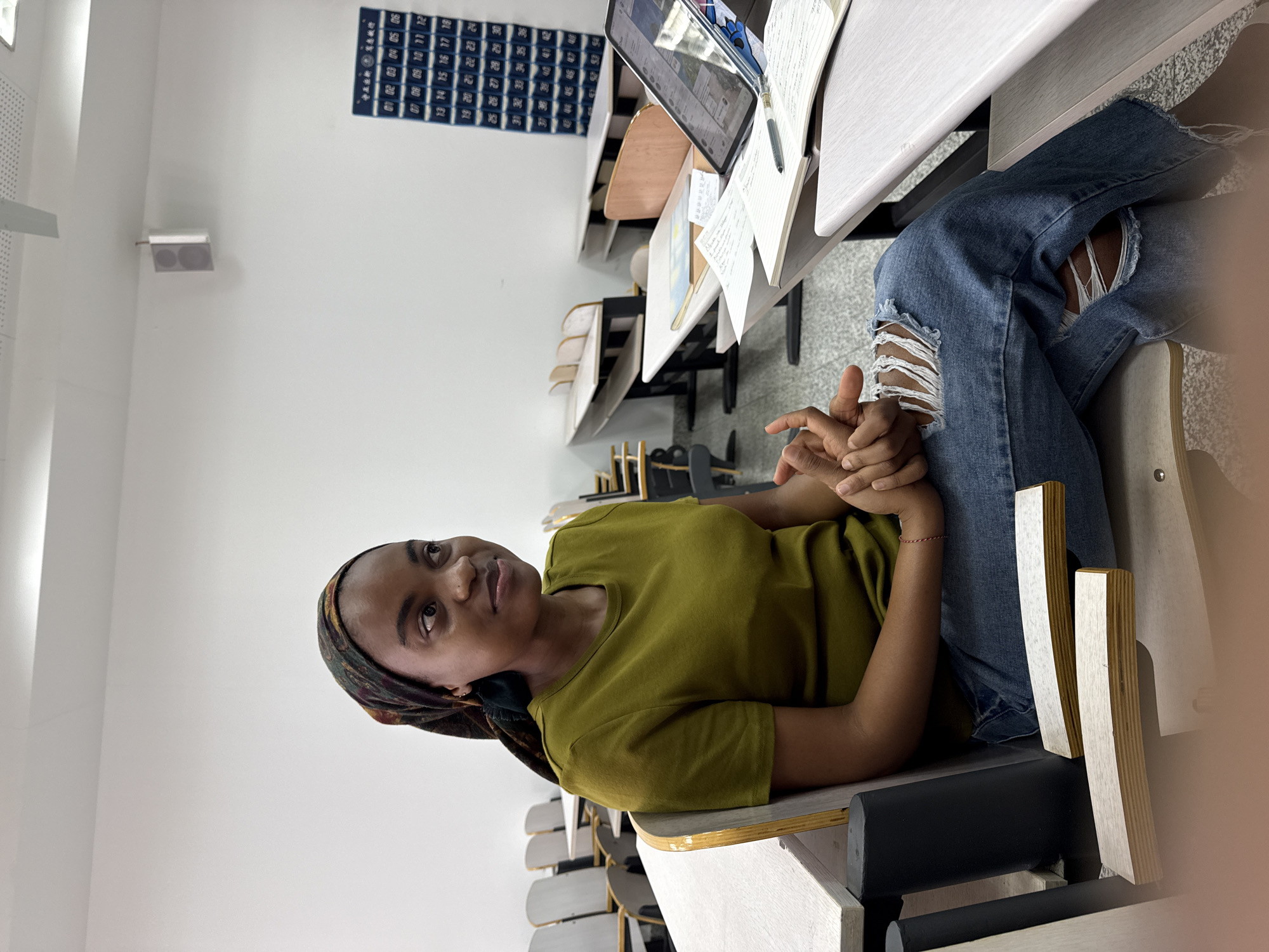 Here is a detailed description of the image:
**Scene Overview:**
The image depicts a young woman sitting in a classroom setting. She is seated at a student desk among rows of similar desks. Books and a tablet are on the desk in front of her.
**People:**
A young woman is the main subject. She has dark skin and is wearing a headscarf and an olive-green t-shirt. She is wearing ripped denim jeans. Her hands are clasped together.
**Objects:**
* Student desks and chairs
* Books
* Tablet
* Writing utensil.
* Bulletin board with numbers on it.
**Environment:**
The scene is a classroom with rows of desks. The walls are plain white.
**Inferred Location:**
Based on the information available, the image was taken in the city of Daxing District, China. The bulletin board may be calendar days.