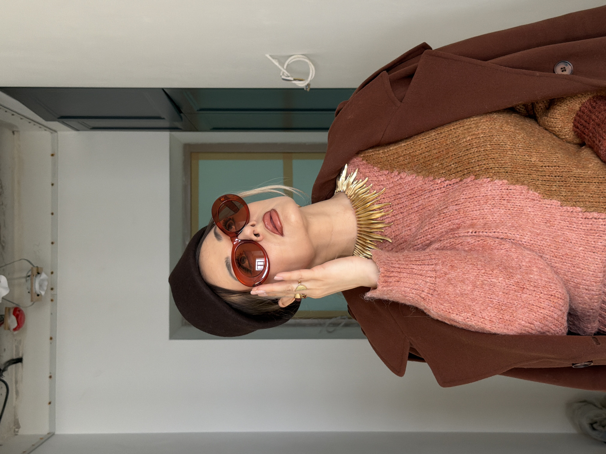 A woman is oriented sideways in an indoor setting. She is wearing a dark brown beret, round red sunglasses, a striped sweater of pink and brown hues, and a brown coat. She is also wearing a gold collar necklace with pointed spikes. Her right hand is raised to her face, displaying a gold ring. The background includes a window with a light green interior, dark gray cabinets, and a white wall. In the upper left corner, there is an unfinished section of the wall. The location cannot be determined from the image.