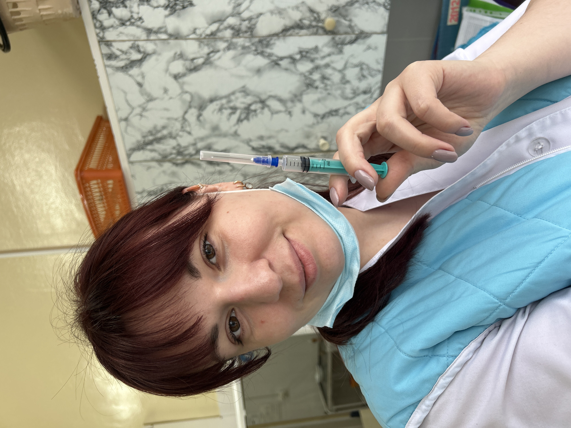 A woman in a blue and white medical coat holds a syringe with a needle in her right hand, while wearing a surgical mask pulled down under her chin. She has brown eyes and maroon hair with bangs. The background includes a wall with a faux marble pattern, a cream-colored wall section, and a glimpse of a bright orange basket. The context of Фоки, Russia is not evident from the image.