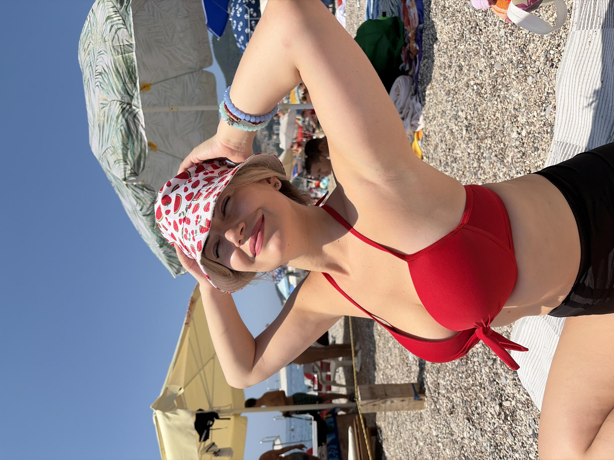 A young woman with blonde hair and a strawberry-patterned bucket hat is sitting on a beach in Budva, Montenegro. She is wearing a red bikini top and black bottoms. Her hands are on top of her head, and her eyes are closed as she smiles. In the background, several beach umbrellas, sunbathers, and the sea are visible. The ground is covered in pebbles.