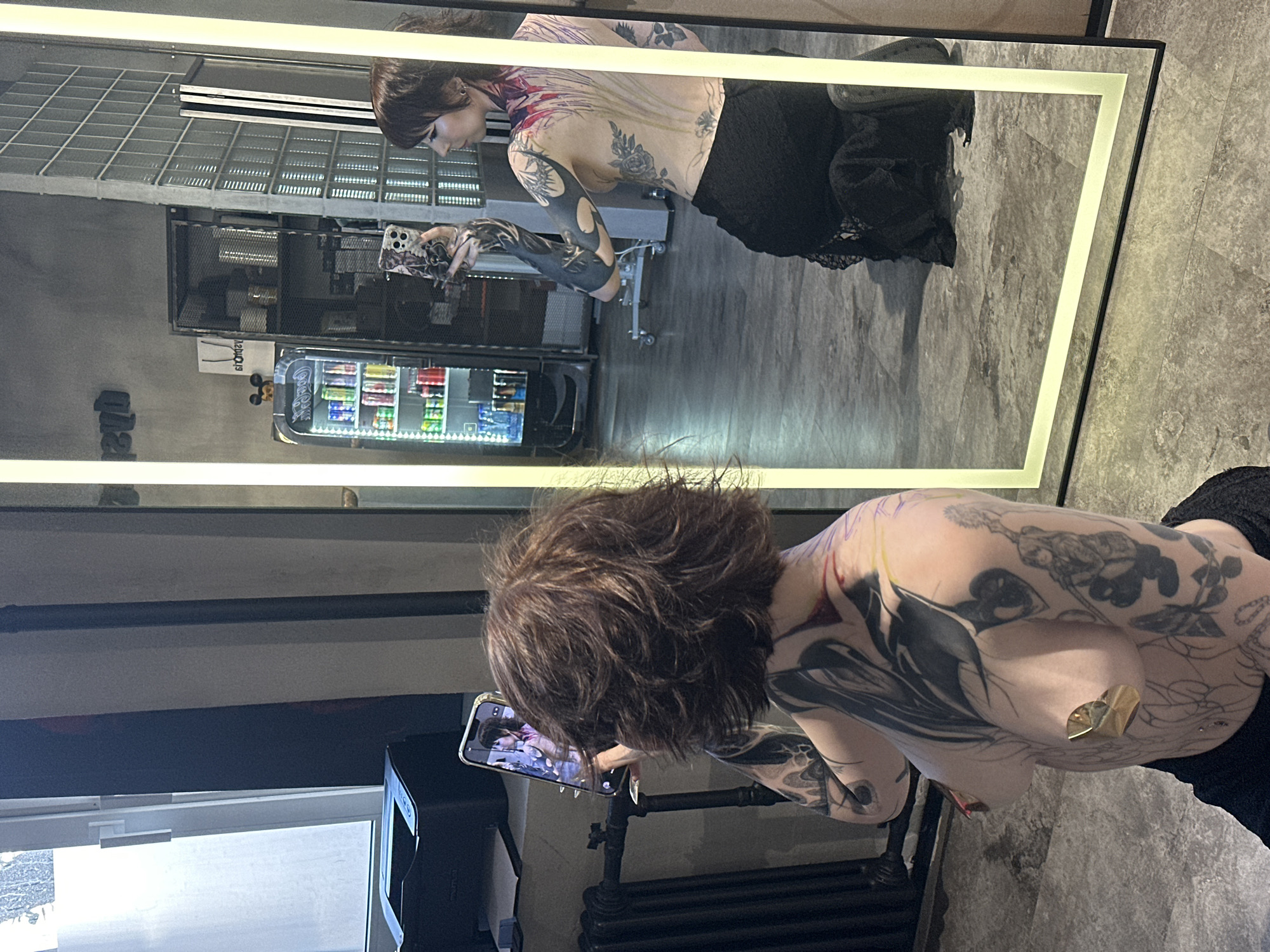 A tattooed person is taking a picture of themself in a mirror. In the reflection, a vending machine is visible.