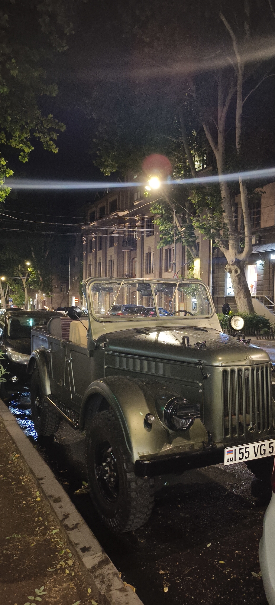 An antique olive green convertible Jeep is parked on a wet, tree-lined city street at night. The Jeep has a spare tire mounted on the hood, a chrome grille, and a license plate that reads "155 VG 5". The street is lit by streetlights and lights from a multi-story building in the background. There are other cars parked behind the Jeep, and the ground is covered in puddles and fallen leaves. The overall scene suggests a vintage aesthetic in an urban environment.