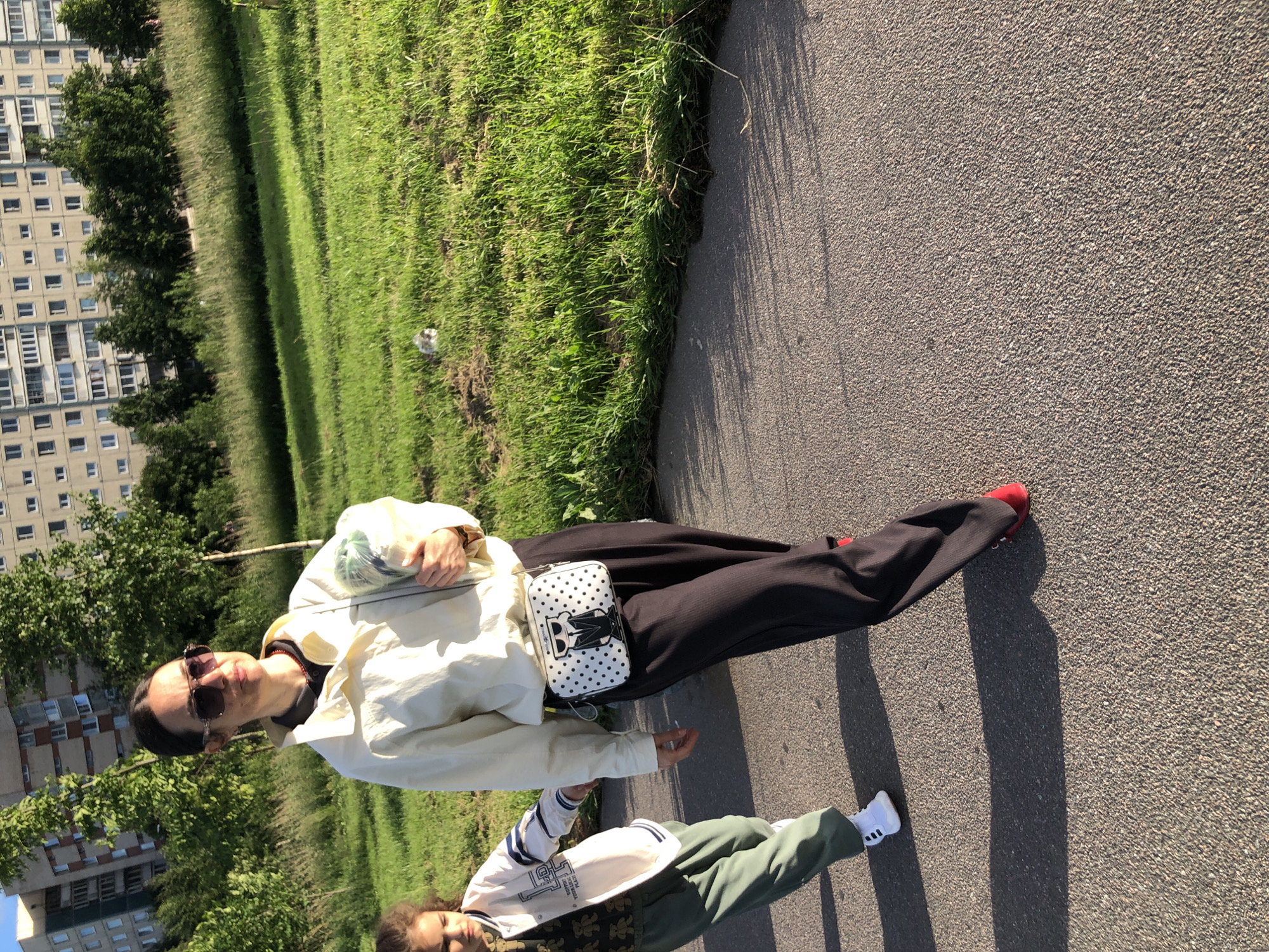 A woman wearing sunglasses, a light-colored jacket, and dark pants walks on an asphalt path. She is carrying a bundle of items in her right arm and has a small crossbody bag with a cartoon design. To her left, a child in a white jacket and green pants walks alongside her, looking down. The background features a grassy area with trees and a multi-story building. The scene is set in Saint Petersburg, Russia, during daylight hours.