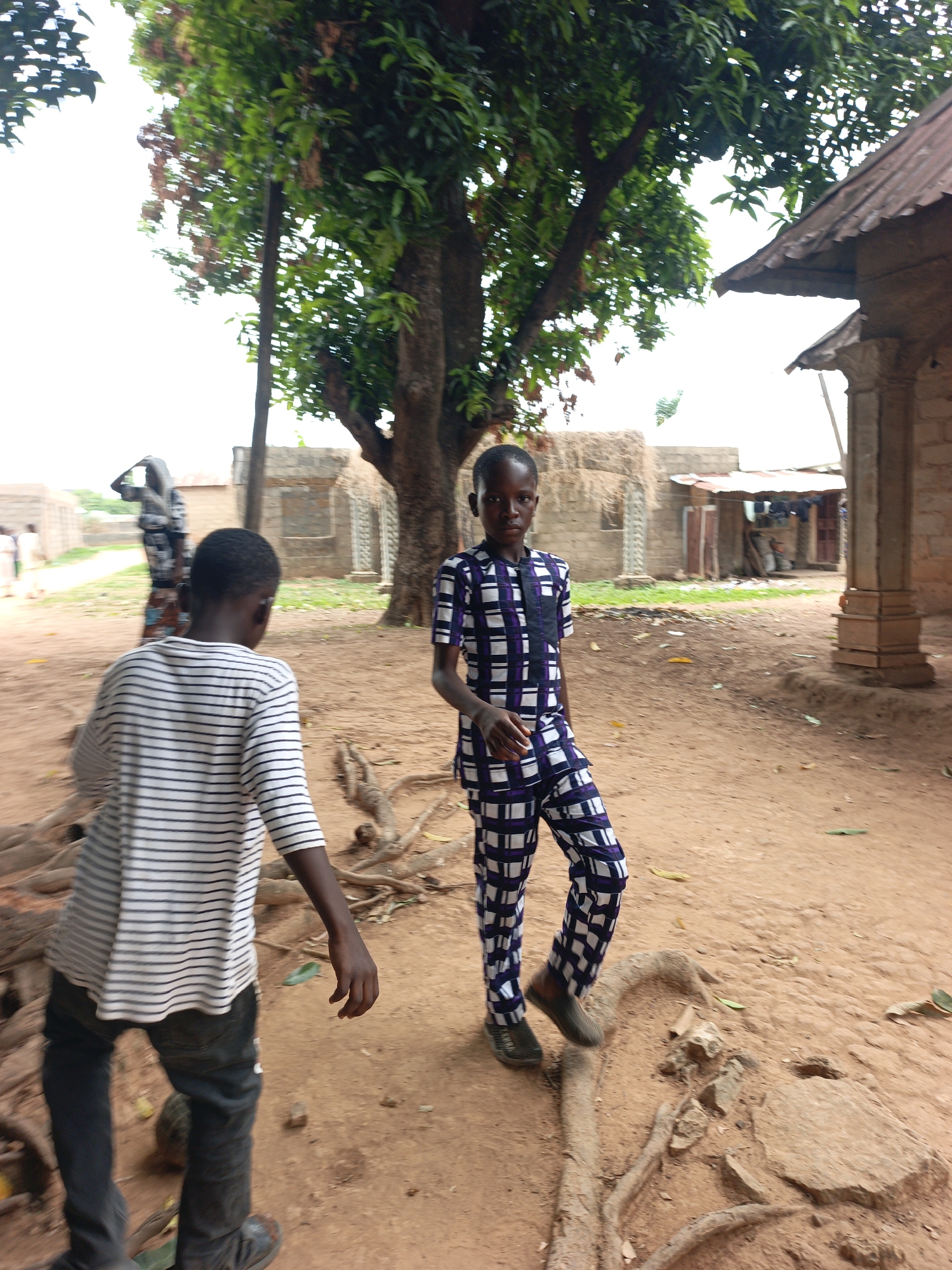 Two boys stand on a dirt lot in Akwanga, Nigeria. The boy in the foreground wears a white and black striped shirt and dark jeans. He appears to be walking to the left. A second boy stands facing the viewer wearing a blue and white checkered patterned outfit. A large tree is behind him. Buildings of brick and straw are scattered in the background. The ground is dirt, with exposed tree roots.
