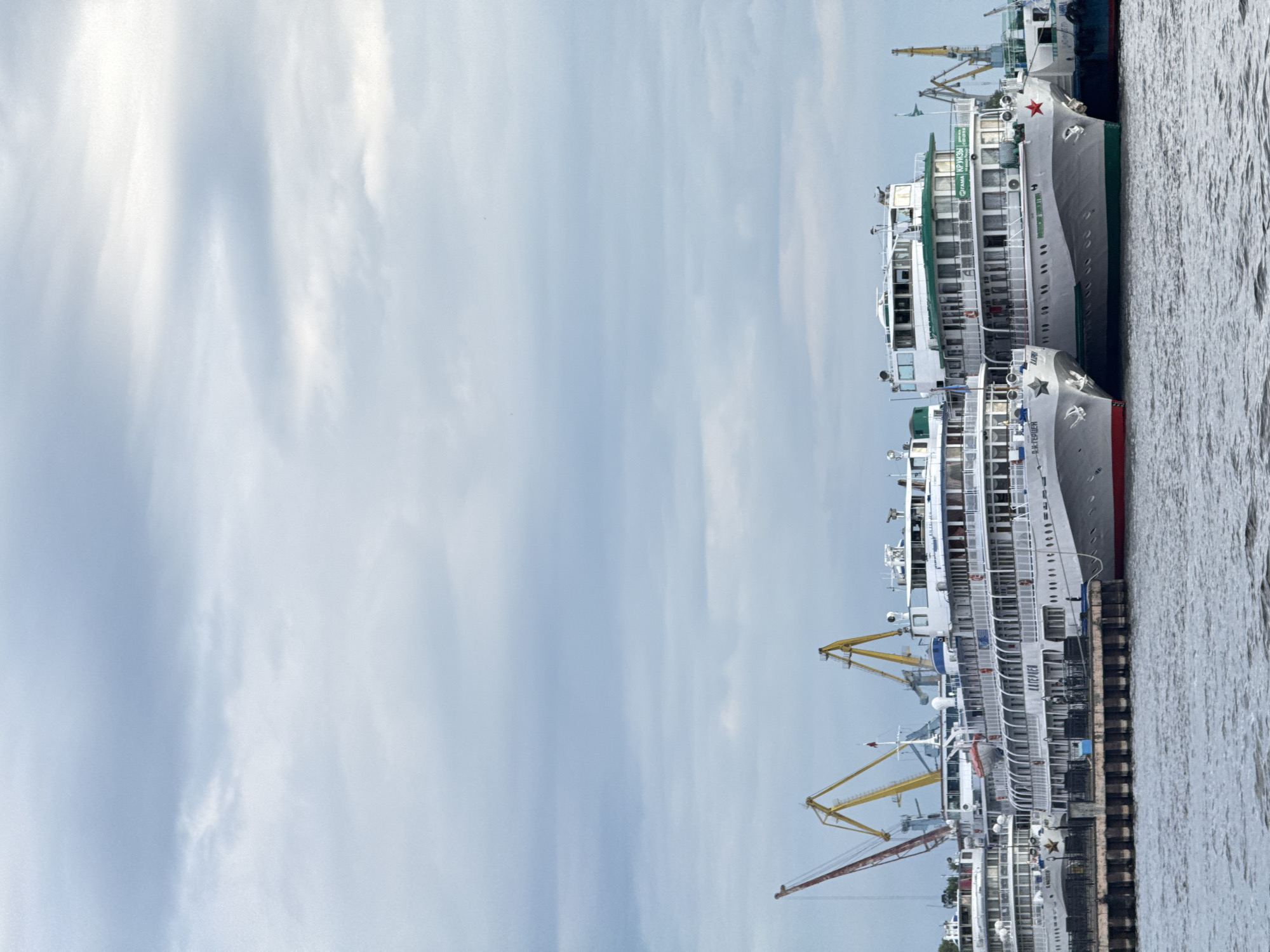 The image depicts a waterfront scene in Kazan, Russia, featuring several large passenger ships docked at a pier. The ships are white with green accents and have multiple decks. Two prominent vessels in the foreground display Cyrillic lettering; one reads "КАТЕРИЯ" and the other appears to say "КАПИТАН". The water in front of the ships is choppy. In the background, several large yellow cranes are visible, suggesting a port or industrial area. The sky is overcast with a mix of light and dark clouds, creating a diffused light. The overall impression is one of a working port with passenger vessels preparing for or having completed a journey.