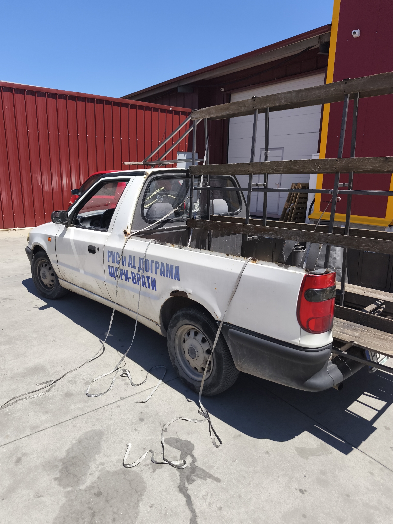 A white pickup truck is parked on a concrete surface outdoors. The truck has a red cab roof and blue lettering on its side that reads "РУСИ И АЛ ДОГРАМА ЩОРПИ-ВРАТИ". A metal ladder is loaded in the truck bed and secured with straps that also lie on the ground. The truck is parked in front of a red corrugated metal building with a yellow stripe and a portion of a dark red building with a white garage door visible in the background. The sky is clear and blue. The scene appears to be in an industrial or commercial area.