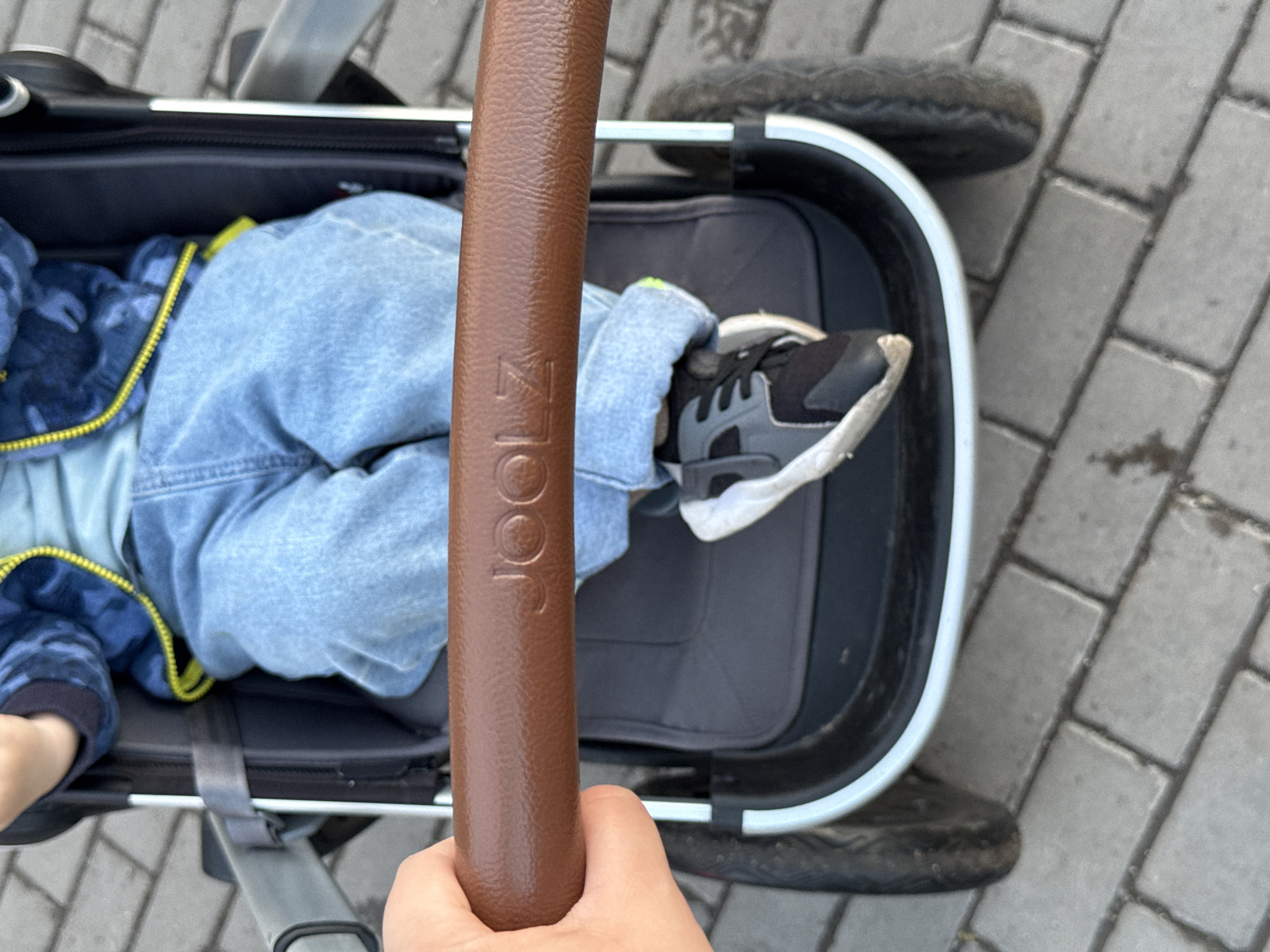 The image shows a close-up view of a child sitting in a stroller. The child is wearing a blue denim jacket with a yellow zipper and light blue jeans. The stroller has a brown leather handle with the brand "JOOLZ" embossed on it. The child's feet are visible, wearing black and grey sneakers. The stroller is on a paved sidewalk made of grey rectangular bricks. The background is slightly blurred, showing more of the brick pavement and the stroller's wheel. The scene appears to be outdoors, in a city environment. There is no indication that the image was taken in Krasnoyarsk, Russia.