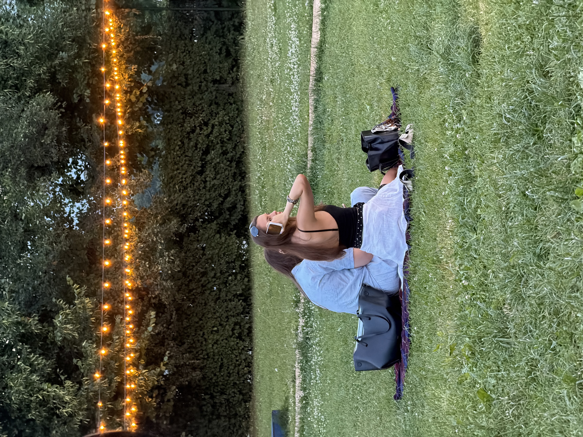 Two individuals are seated on a blanket on a grassy lawn, facing away from the camera. The person in the foreground, on the right, is wearing a black tank top and a white skirt, and is holding a mobile phone to their ear. They have sunglasses perched on their head. The other individual, on the left, is wearing a light blue shirt and pants. In front of them, on the blanket, are two large black tote bags and a pair of sandals. The background features a dense line of green trees and bushes, with a strand of warm-toned string lights strung across the upper portion of the frame, casting a glow. The ground is covered in green grass with scattered small white flowers. The scene appears to be an outdoor public space, possibly a park, in Moscow, Russia, during the evening.
