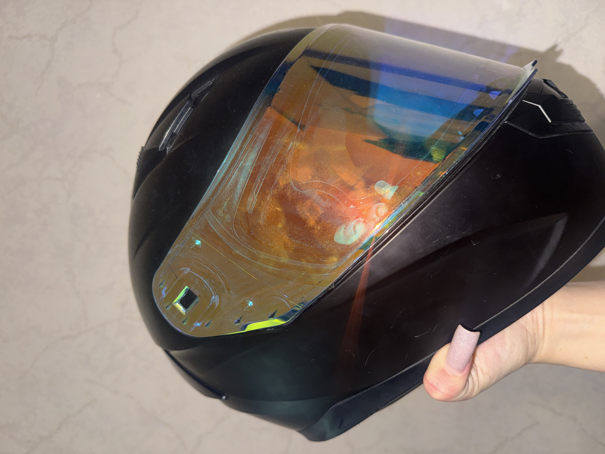A black motorcycle helmet with a reflective, iridescent visor is held in a person's left hand. The visor displays a spectrum of orange, yellow, blue, and green colors due to its reflective coating. The helmet is black and appears to be made of plastic. The hand holding the helmet has a light pink nail polish on the fingernail of the thumb and is positioned on the lower left side of the frame. The background is a plain, light-colored wall with a subtle textured pattern. The image does not provide any information about the location in Bishkek City, Kyrgyzstan.