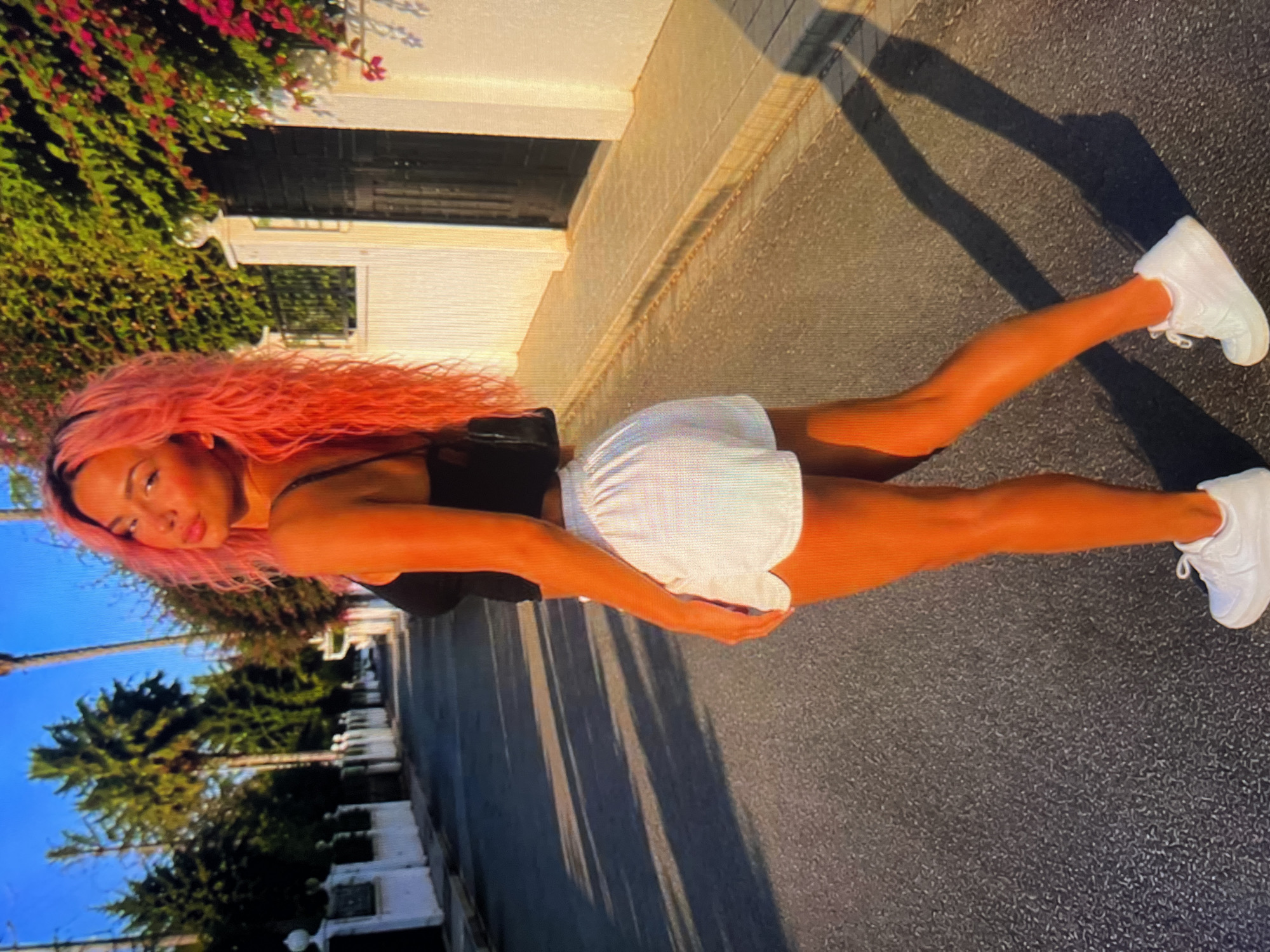 A young woman with vibrant pink hair stands on an asphalt street, looking over her shoulder towards the camera. She is wearing a black tank top and white shorts, with white sneakers on her feet. The sun casts long shadows across the street from the palm trees and white-fenced houses lining the road. The scene is illuminated by the warm glow of what appears to be late afternoon or early evening sunlight, highlighting the woman's tanned skin.