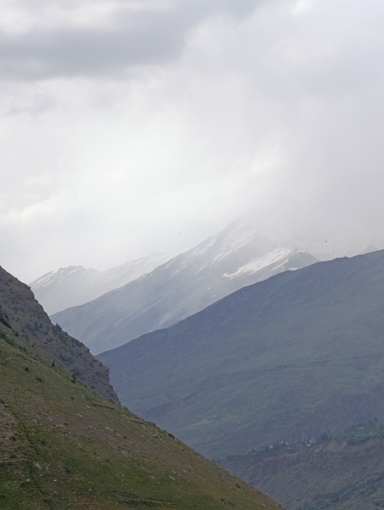 The image depicts a mountainous landscape under a cloudy sky, with several layers of mountains receding into the distance. The foreground features a steep, grassy slope on the left side of the frame, with rocky outcrops and sparse vegetation. Behind this, a darker, more densely vegetated mountain slopes downward. Further in the background, several more mountain ranges are visible, some with patches of snow or ice near their peaks. The atmosphere is hazy, suggesting mist or fog, especially in the mid-ground and background, obscuring details of the distant terrain. In the far distance, a small cluster of buildings, possibly a village, is visible nestled in a valley. The overall scene conveys a sense of natural grandeur and isolation. The provided information states the location is Tandi, India, though specific landmarks or details within Tandi are not discernible from the image alone.