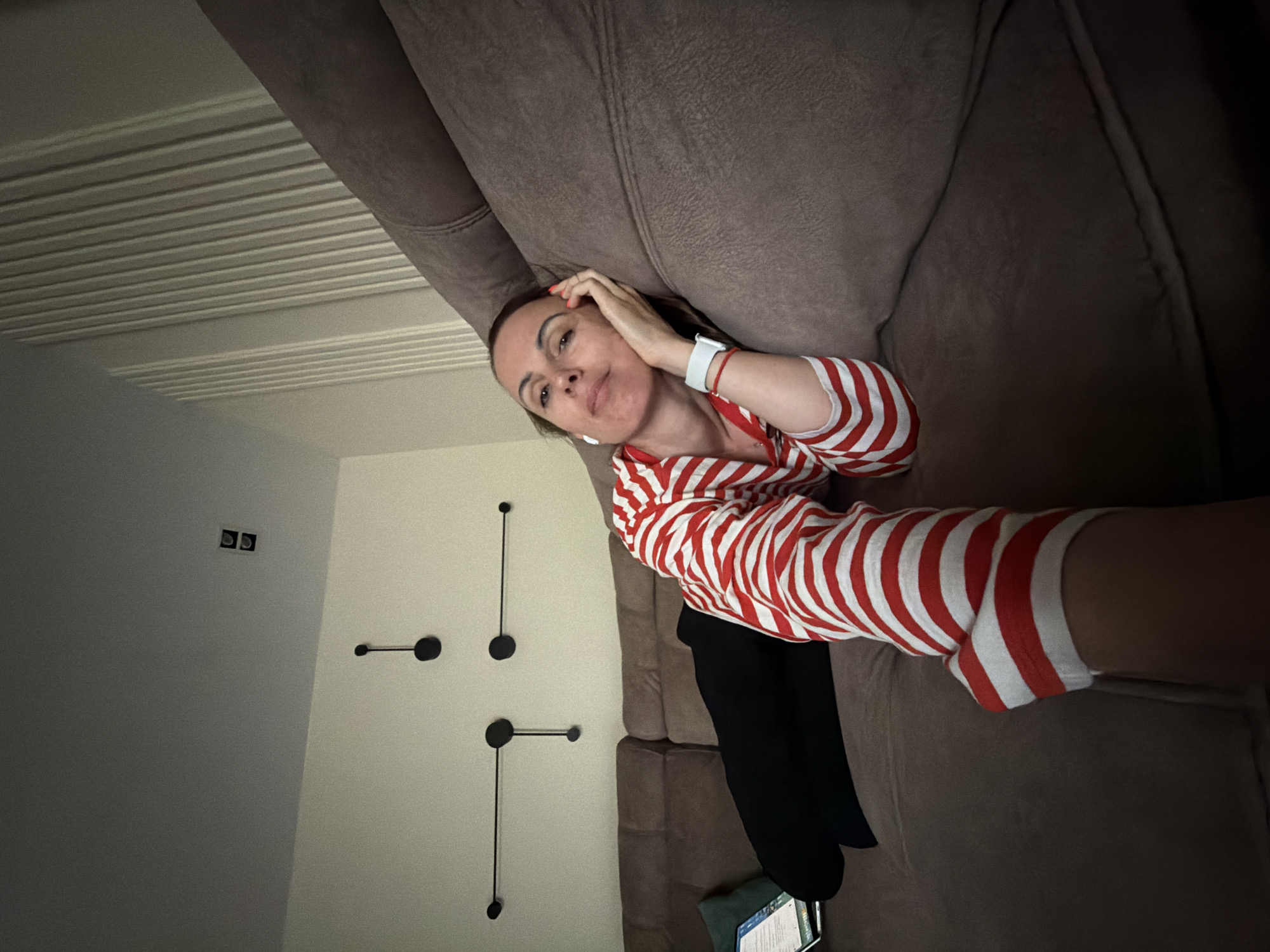 A woman is depicted lying on her side on a beige-brown fabric couch in an indoor setting in Birobidzhan, Russia. She is wearing a red and white striped long-sleeved top and dark-colored pants. A white smartwatch is visible on her left wrist, and she has an earbud in her right ear. Her left hand is casually placed near her head, with red-painted fingernails. She looks towards the camera with a neutral, slightly pensive expression.
The background consists of light-colored walls. On one wall, several modern, abstract black wall decorations are arranged, featuring circular elements connected by thin lines. On the ceiling or an upper wall, distinct horizontal shadow lines are visible, suggesting light filtering through blinds or a textured surface. Near the woman's lower body, a digital device, possibly a smartphone or tablet, is resting on the floor, but any text on its screen is illegible. Further into the room, black square shapes are noticeable on a wall, potentially indicating fixtures or vents. The lighting appears dim but comfortable, contributing to a relaxed ambiance.