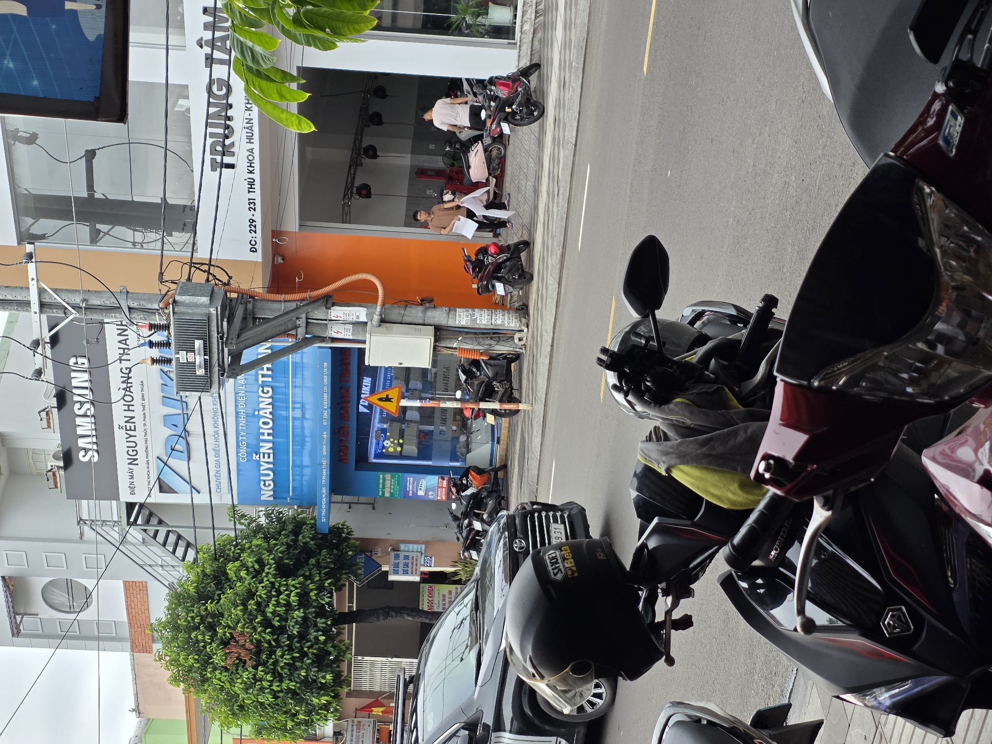 The image depicts a street scene in Phu Thuy Ward, Vietnam, filled with parked motorcycles and a few pedestrians in front of businesses. The focus is on the street and the parked motorcycles occupying the foreground. On the left side of the image, there is a building with signage displaying "SAMSUNG," "DAIKIN," and "NGUYỄN HOÀNG THANH." A tree partially obscures the building. Further down the street, there's a building with an orange facade and signage displaying "TRUNG TÂM." Several people are seen standing and sitting near the entrance of that building. The street is paved and marked with a yellow line. A car is also visible, parked between the motorcycles. A yellow triangular traffic sign is mounted on a pole, partially obscured by the tree.