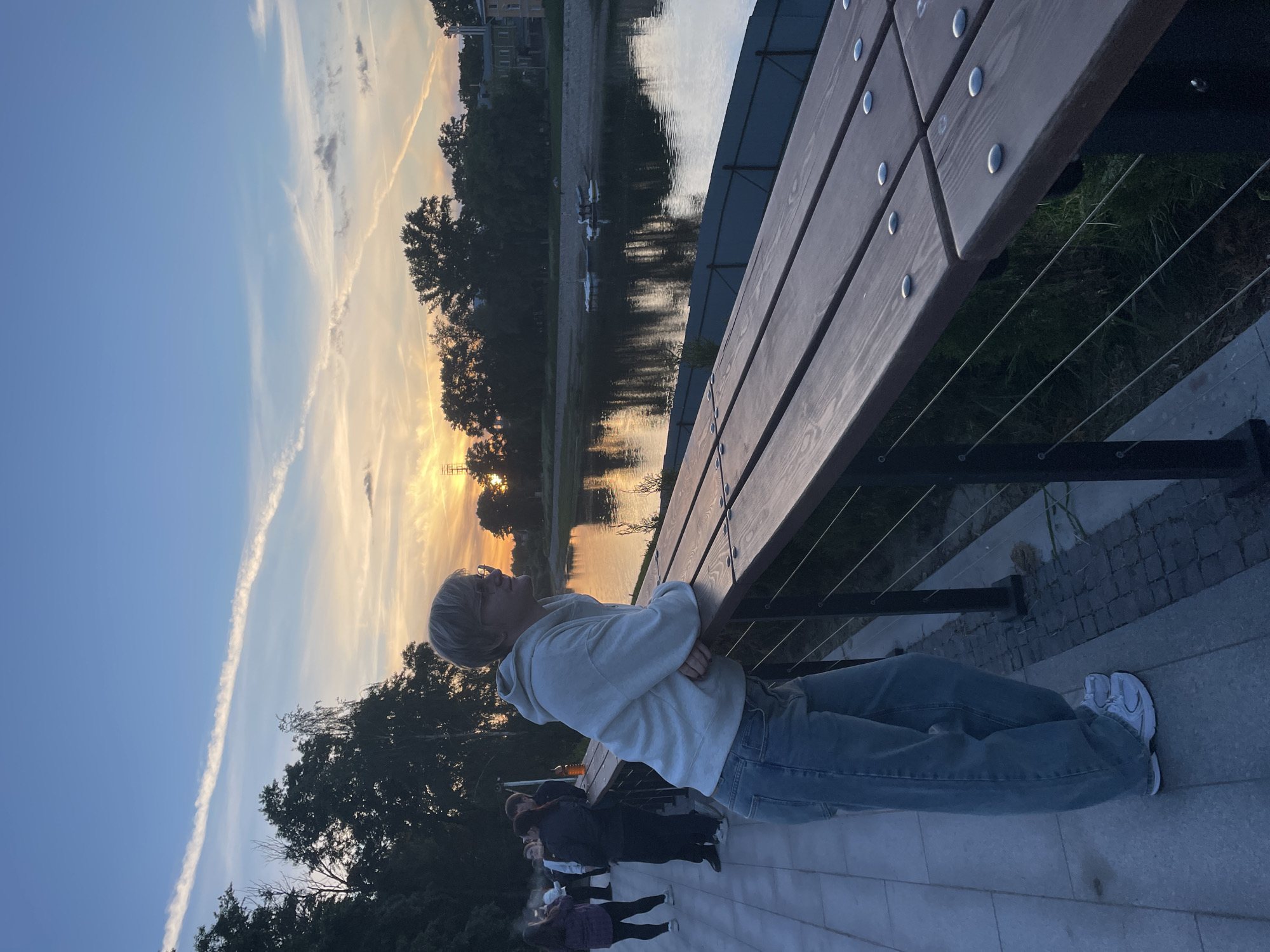 This image captures an outdoor scene at sunset by a body of water in Vologda, Russia. The primary subject is a person, likely male, with short blonde hair and glasses, wearing a light grey hooded sweatshirt, blue jeans, and white sneakers. They are leaning with their arms crossed on a wooden bench, gazing contemplatively towards the setting sun.
The scene unfolds during dusk, with the sky displaying a beautiful gradient from clear blue on the left to warm yellows and oranges near the horizon, indicating a sunset. Wispy clouds and a prominent, long contrail streak across the sky. The warm light of the setting sun is reflected on the calm surface of a river or lake that occupies the middle ground of the image. On the distant bank, a line of dark trees and some indistinct buildings are visible. Two small, light-colored boats are beached near the water's edge on the far side.
In the immediate foreground, a sturdy wooden bench with visible metal bolts runs diagonally, partially obscuring a railing made of metal posts and thin wires. Several other people, out of focus and casually dressed, are visible in the background on the same side as the main subject, suggesting a public promenade or park area. The overall atmosphere is tranquil and relaxed, highlighting a quiet evening moment. There is no visible text in the image.