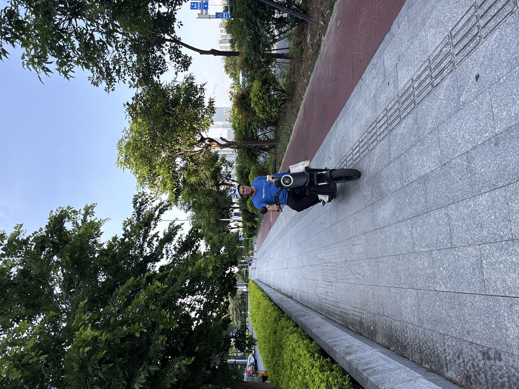 In the Haizhu District of China, a man wearing a blue t-shirt with "Trip.com" printed on it and a white helmet rides a black electric scooter along a paved sidewalk. The scooter has a white bag attached to the front. The sidewalk is bordered by a red bike lane on the right and a raised planter with green bushes on the left. Beyond the planter, cars are parked along a street. Trees line the sidewalk and bike lane, with a clear blue sky above. In the background, tall buildings and more trees are visible, suggesting an urban environment. The lighting indicates it is daytime.
