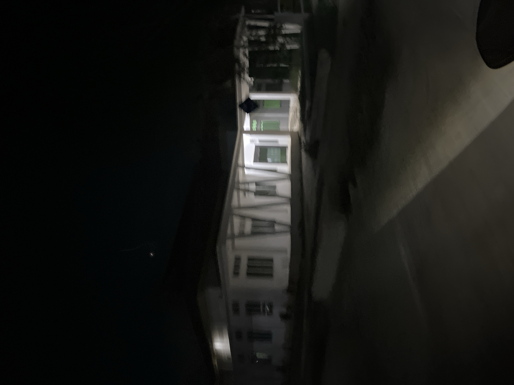 The image depicts an outdoor scene at night in Bundar, Indonesia. The primary subject is a white, multi-story building with several windows and doors. Some of the windows appear to be illuminated with a green light, suggesting they are interior lights. The building seems to be under construction or recently completed, with exposed structural elements visible.
A person, wearing dark clothing, is standing on the ground in front of the building, partially obscured by the darkness. The ground in front of the building is paved, possibly a sidewalk or driveway. To the right of the building, there's a darker area that might be a road or an unlit pathway.
The overall lighting is dim, characteristic of nighttime. There is a faint light source visible in the far left of the image, possibly a distant streetlamp or a celestial body. The image has a slightly blurry quality, suggesting motion or low light conditions during capture. No text is visible in the image. The scene appears calm and quiet, with no indication of any specific event or strong emotions.