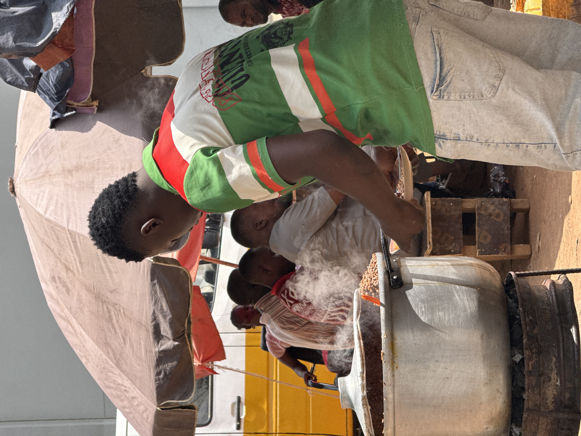 In Abuja, Nigeria, a group of people are gathered around a large metal pot, presumably cooking food. One person, wearing a green, white, and red striped shirt and light blue jeans, is leaning over the pot, perhaps stirring or observing its contents. Smoke rises from the pot, suggesting it is heated by a fire beneath. An umbrella provides shade for the individuals. A yellow truck is visible in the background.