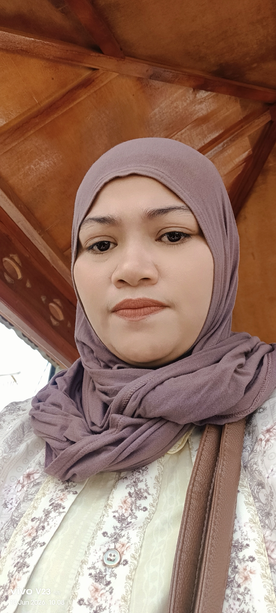 A portrait of a woman wearing a light purple hijab and a floral patterned shirt. She is looking directly at the camera with a neutral expression. Behind her, a wooden ceiling structure is visible. The image also shows a portion of a brown bag strap on the right side of the frame. The bottom of the image contains a watermark with the text "vivo V23 5G 13 Jun 2026, 10.08". The setting appears to be outdoors in a shaded area with wooden architectural elements.