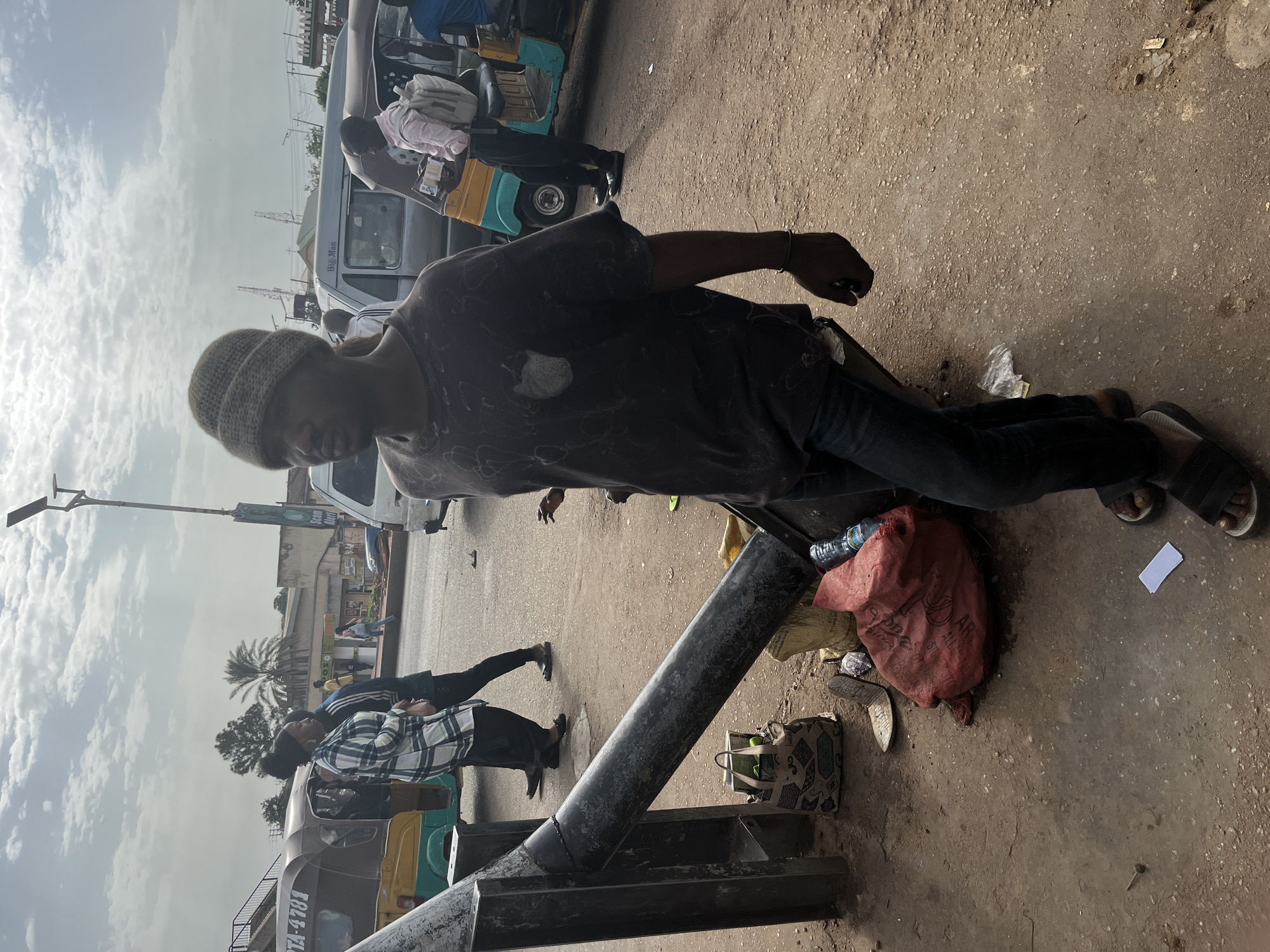 In Gura Top, Nigeria, a man wearing a grey knitted beanie and a dark t-shirt with a faded pattern walks past a black metal pole. He is dressed in blue jeans and black sandals. Next to the pole, a large red sack, a patterned tote bag with a green object inside, and a yellow bag are on the ground. In the background, several auto-rickshaws and vans are parked on a dirt road. Two women in a checkered shirt and a person in a light pink shirt with a backpack are visible. The sky is overcast.