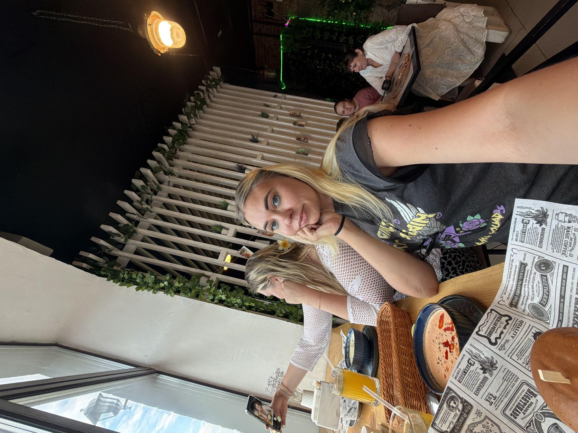 In a restaurant in Chaikovsky, Russia, two women are taking a selfie at a table by a large window. The woman in the foreground is wearing a dark t-shirt with a graphic print and has blonde hair with blue eyeshadow. The woman behind her is wearing a white mesh top and has blonde hair. In front of them are bowls of soup, a glass of orange juice, and a burger. The restaurant has a modern interior with white slatted walls and green ivy decorations. In the background, two other women are seated at a table, one wearing a pink shirt and the other a white lace top, with a pizza on the table. The lighting is warm and inviting, with a hanging lamp visible in the upper right corner of the image.
