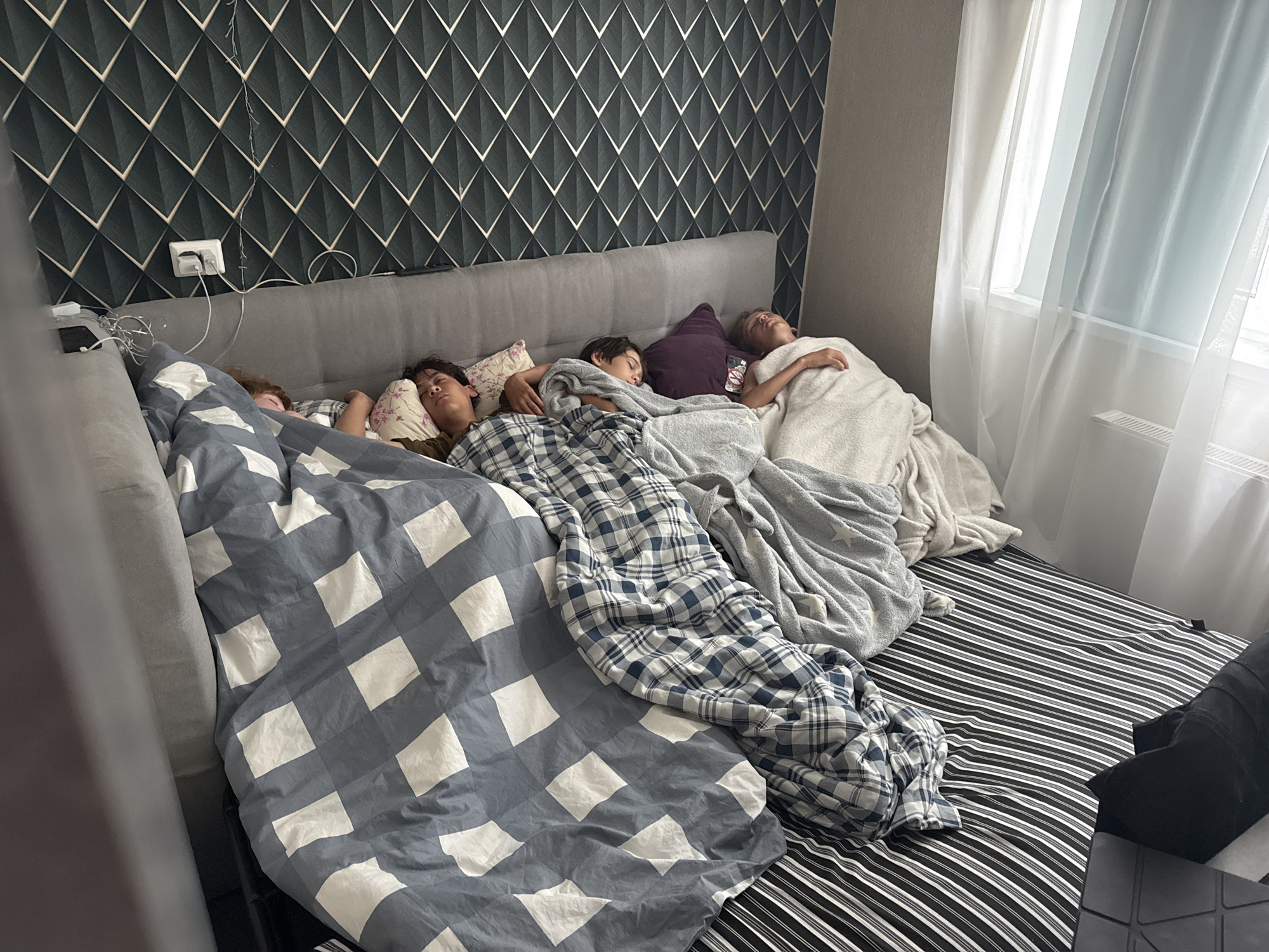 Four young individuals are sleeping on a large bed in a room. The bed is covered with various blankets and pillows. The background features a patterned wallpaper and a window with sheer curtains. A power outlet with a charger plugged in is visible on the wall. The scene appears to be a bedroom.