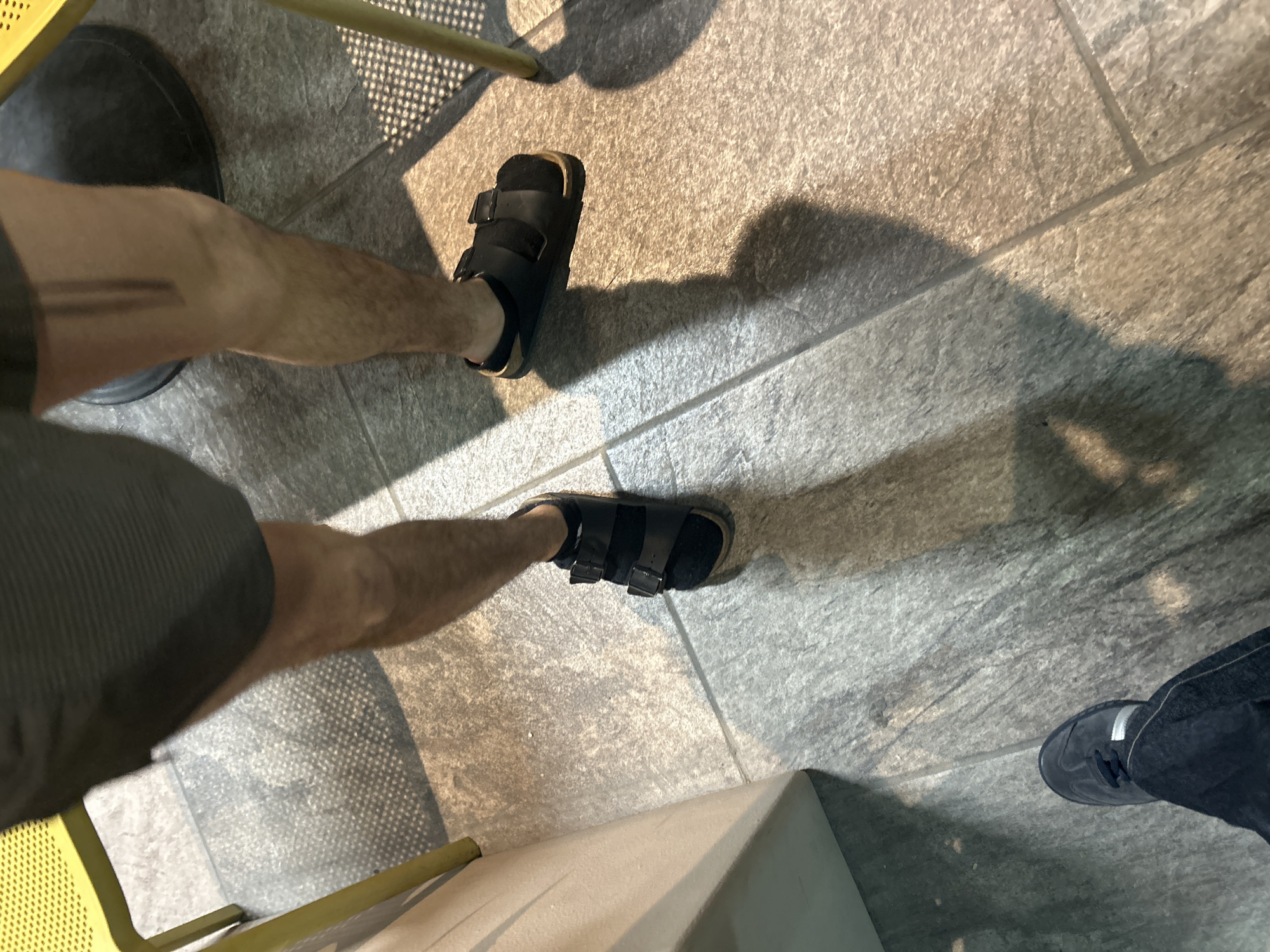 A high-angle shot captures a person sitting with their legs angled apart. The individual is wearing black sandals with adjustable straps. One foot is positioned slightly above the other on a stone tiled floor. Part of the person’s leg is visible, wearing a dark colored garment. The lower legs are bare. The feet appear to be on opposite sides of a small yellow chair. Another person’s leg and sneaker-clad foot are visible in the lower right corner of the shot. The setting appears to be an indoor space with the focus on the floor and the legs of the subjects.