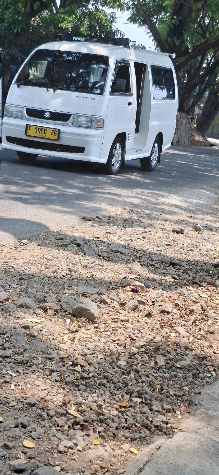A white Suzuki Carry 1.3 van is parked on the side of a paved road. The van's front license plate reads "F 1950 ZG". The driver's side door of the van is open. The area around the van is covered in gravel, leaves, and some small rocks. Trees with lush green foliage are visible behind the van, and their shadows are cast on the road. The environment appears to be an outdoor scene, possibly in a residential or semi-urban area, consistent with locations in Cisalak, Indonesia. The image captures a still moment, with no active movement depicted.