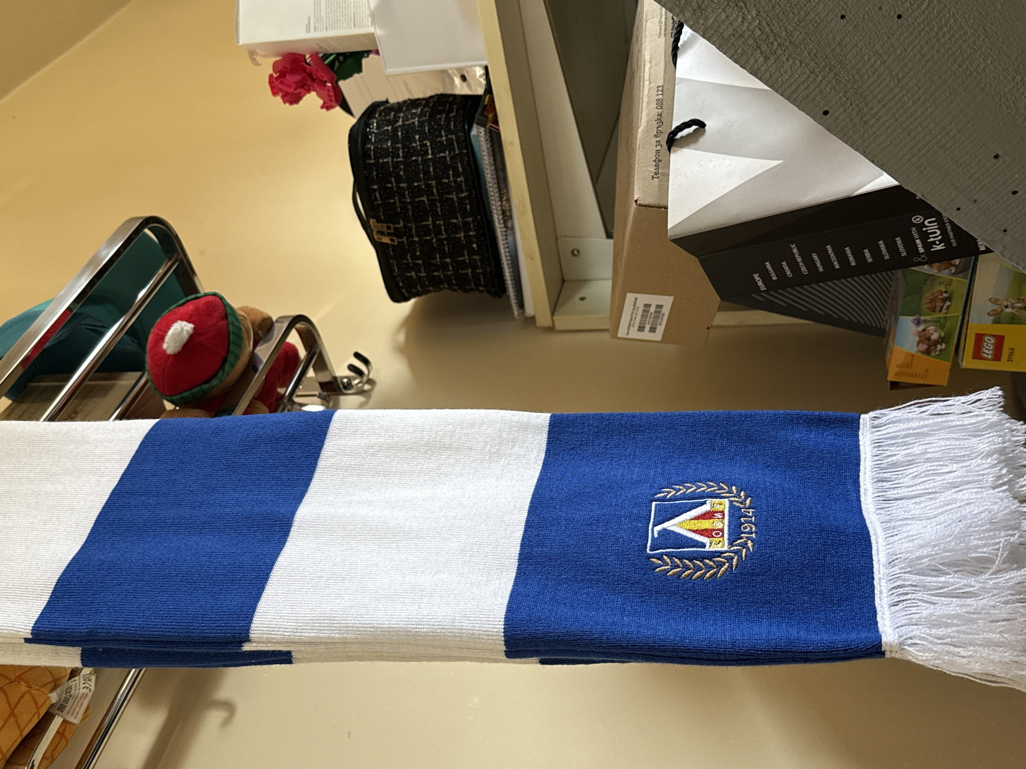 An indoor, static shot captures a collection of objects arranged on shelves and surfaces, presented with a 90-degree counter-clockwise rotation. No people are visible, and no activity is taking place.
The prominent subject in the foreground is a blue and white striped scarf, neatly folded. The blue section of the scarf features an embroidered logo consisting of a stylized 'V' shape, the text "SOFIA" and "1914", all encircled by golden laurel wreaths. One end of the scarf has white fringes.
In the mid-ground, a red and white plush toy, possibly a character with a white pompom, rests on a silver metal rack. Above this, a black textured handbag with a gold clasp sits on a shelf. A red artificial flower is also visible nearby.
The background is cluttered with various boxes and papers. One box clearly displays "LEGO 31162". Another cardboard item features the "k-tuin" logo and a list of European countries including "BULGARIA", "CROATIA", "CZECH REPUBLIC", "HUNGARY", "MACEDONIA", "ROMANIA", "SERBIA", "SLOVAKIA", "SLOVENIA", and "SPAIN". Further text on a piece of paper or cardboard reads "Телефон за връзка: 088 123" (Contact phone: 088 123), which is in Bulgarian. The surfaces are a light yellow/beige, illuminated by artificial indoor lighting. The "SOFIA" text on the scarf and the Bulgarian text and country listing support the given location context of Sofia, Bulgaria.