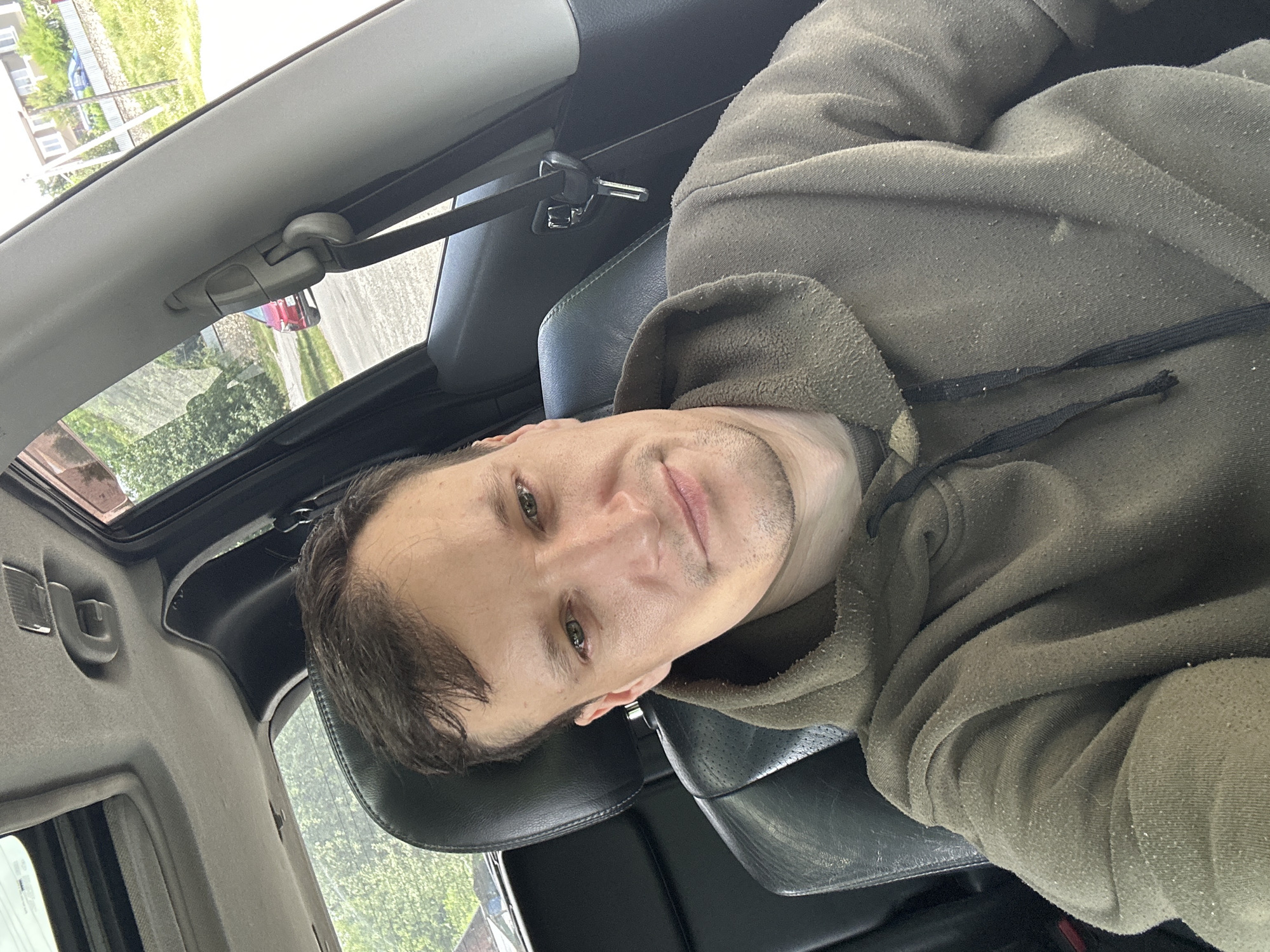 A man with short brown hair and green eyes is taking a selfie inside a car. He is wearing a brown hooded sweatshirt. The car's interior is visible, including the black leather seats, the sunroof, and a seatbelt. Through the car windows, trees and houses can be seen, suggesting an outdoor, possibly rural or suburban, setting. The general appearance of the surroundings implies the location is Makiv, Ukraine, based on the request.