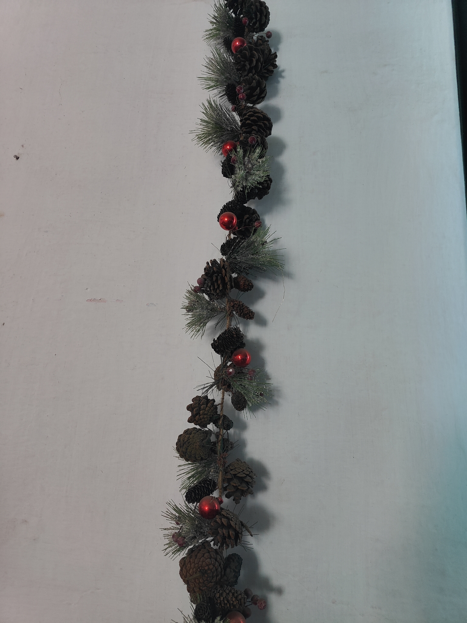 A vertical garland composed of pine cones, artificial pine needles, and small red baubles is displayed against a light-colored, textured wall. The garland hangs from top to bottom on the right side of the frame. The wall appears to be an interior surface, possibly a painted wall in a room. The lighting suggests it is indoors, with some shadow casting on the left side of the garland. There are no people or animals visible in the image. The location of Бобылево, Russia is not determinable from the visual information provided in the image.