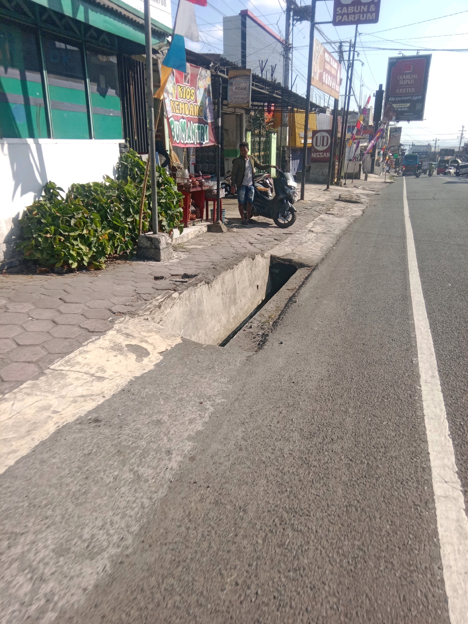 The image depicts a street scene in Kertek, Indonesia, on a sunny day. In the foreground, the asphalt road features a white lane marking. To the left of the road is a concrete drainage ditch, and a paved sidewalk with hexagonal tiles. A young man in a green jacket and denim shorts stands next to a dark-colored scooter parked on the sidewalk. He is leaning against the scooter, with his left hand on the handlebars. Behind him, various businesses are visible, including one with a green facade and large windows, and another with a banner that reads "KIOS KEMBANG ROMANTRI". Further down the street, there are other signs, including one for "SABUN & PARFUM" and a large billboard advertising "DIARUM SUPER KRETEK". Several flags are also visible. The environment appears to be a commercial or semi-urban area with moderate traffic, as indicated by the presence of other vehicles in the distance.