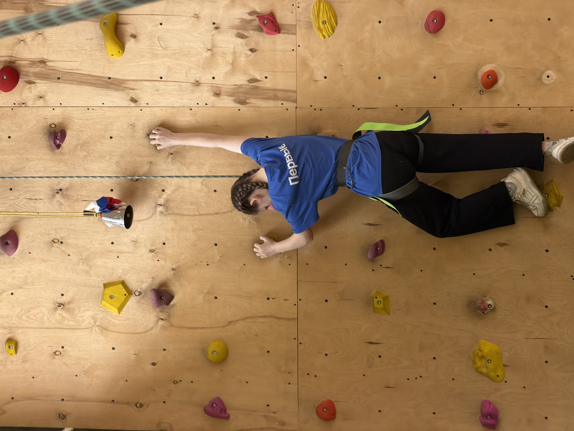 A young person is climbing on an indoor climbing wall. The wall is made of wood and has various colored climbing holds attached. The climber is wearing a blue t-shirt with white Cyrillic lettering "Первый" ("First") on the back, black pants, and white sneakers. They have a green harness and are attached to a rope. The location is likely a climbing gym or recreational facility, possibly in Izhevsk, Russia given the lettering on the shirt.