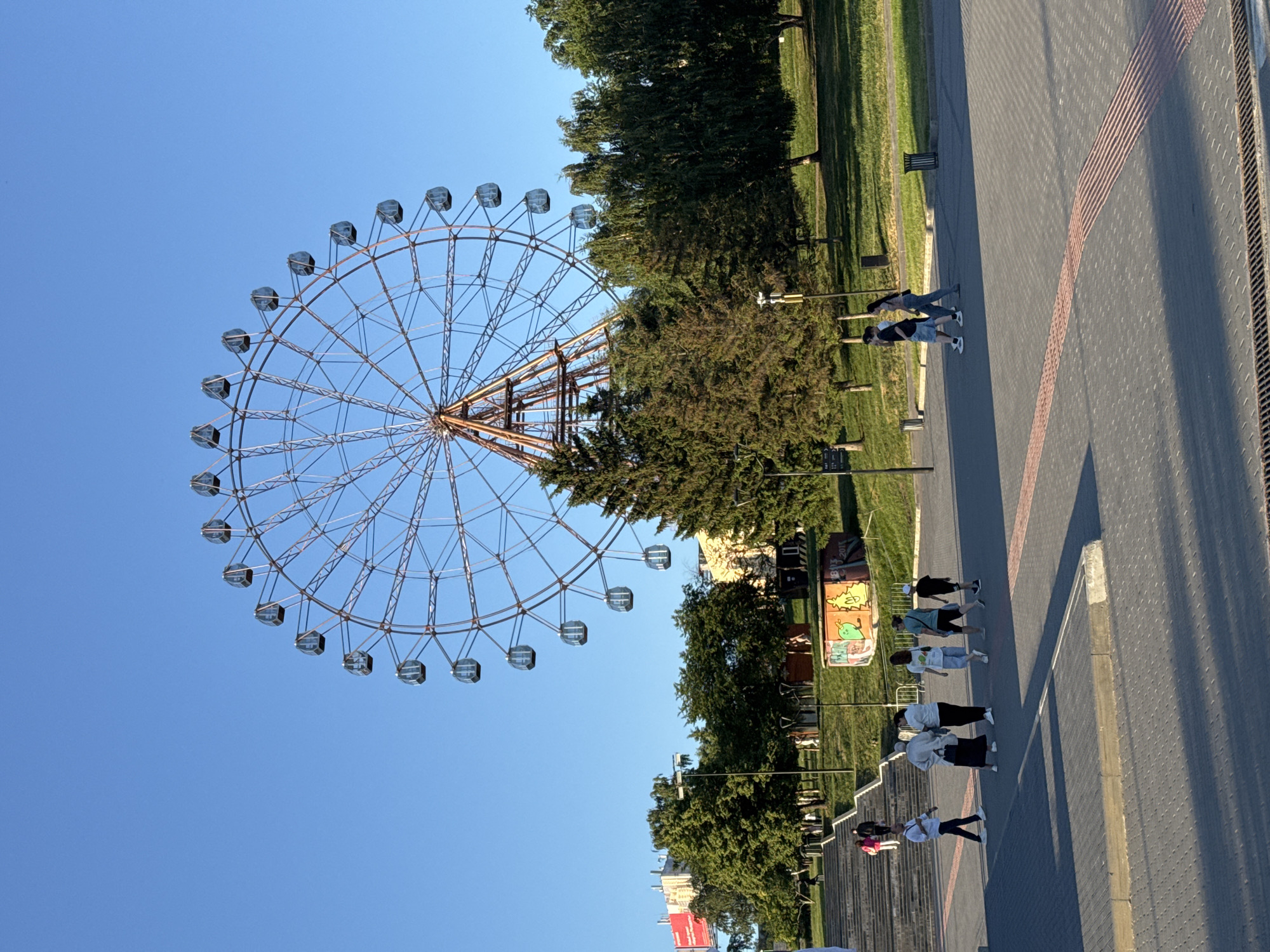 A wide shot shows a Ferris wheel in the background of a park area with trees and a clear blue sky. In the foreground, a group of people are walking on a paved area. The Ferris wheel is a large, metal structure with enclosed cabins. Trees are scattered throughout the park, with some taller pine trees. The paved area is made of gray interlocking stones, with a red tactile paving strip running through it. A set of stairs is visible to the left, leading up a grassy hill. In the distance, buildings are visible, including one with a red billboard. A small, brightly colored structure with graffiti is also present in the park area. The scene appears to be an outdoor public space in Novosibirsk, Russia, likely a park or amusement area, during daylight hours.